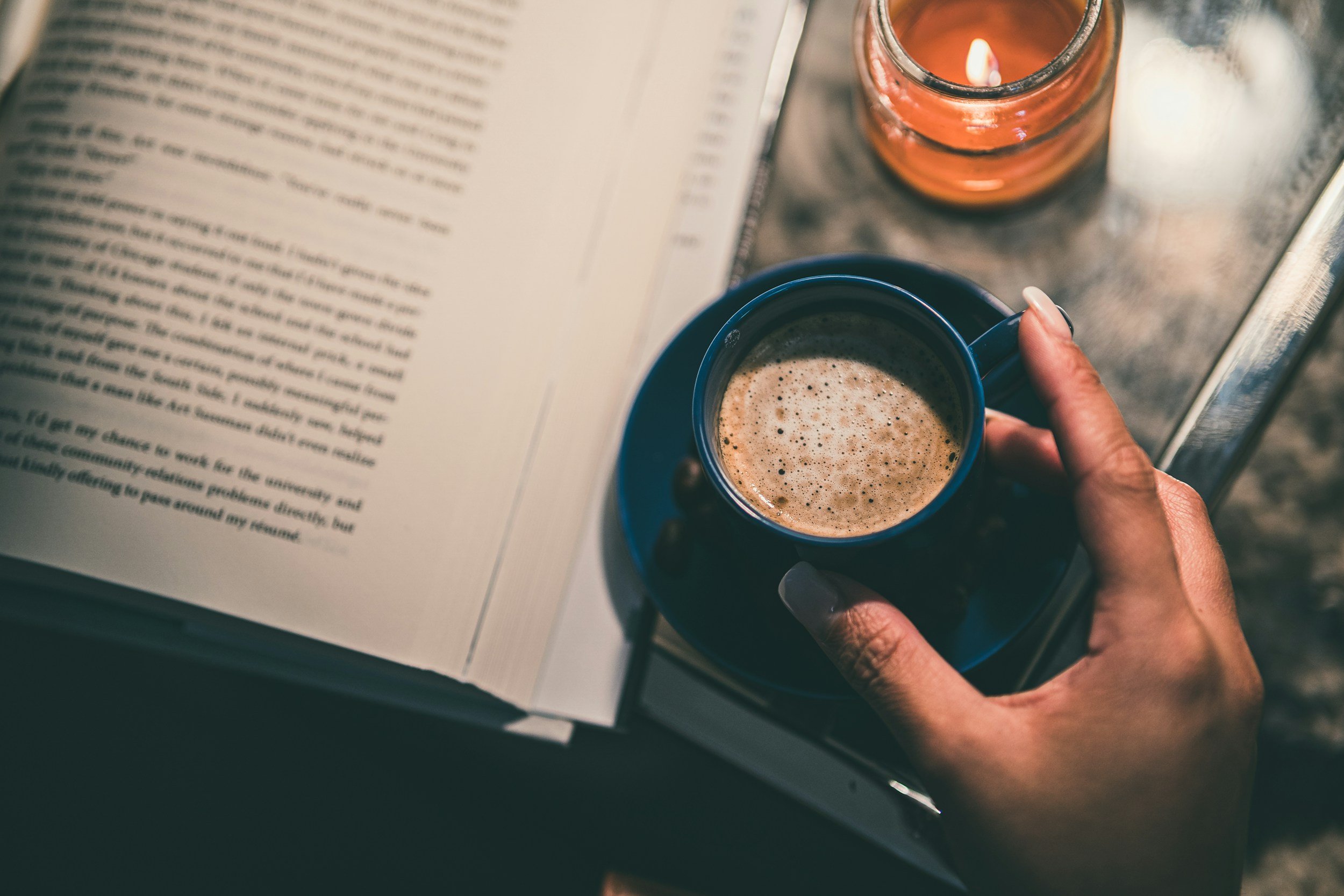 A person holding a blue coffee mug filled with coffee, with an open book and a lit candle on a metal table.