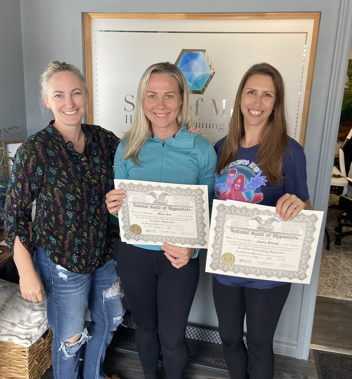Three women smiling, two holding certificates, standing in front of a sign that says 'Self M...'