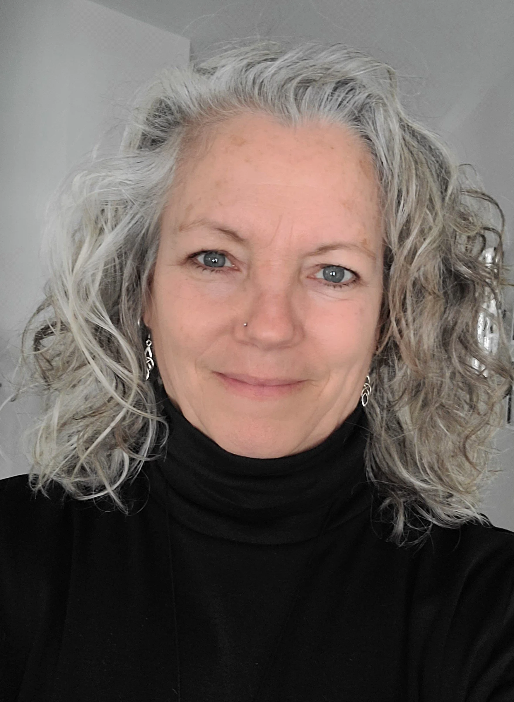 Close-up portrait of a woman with curly gray hair, blue eyes, wearing a black turtleneck and earrings, smiling at the camera.