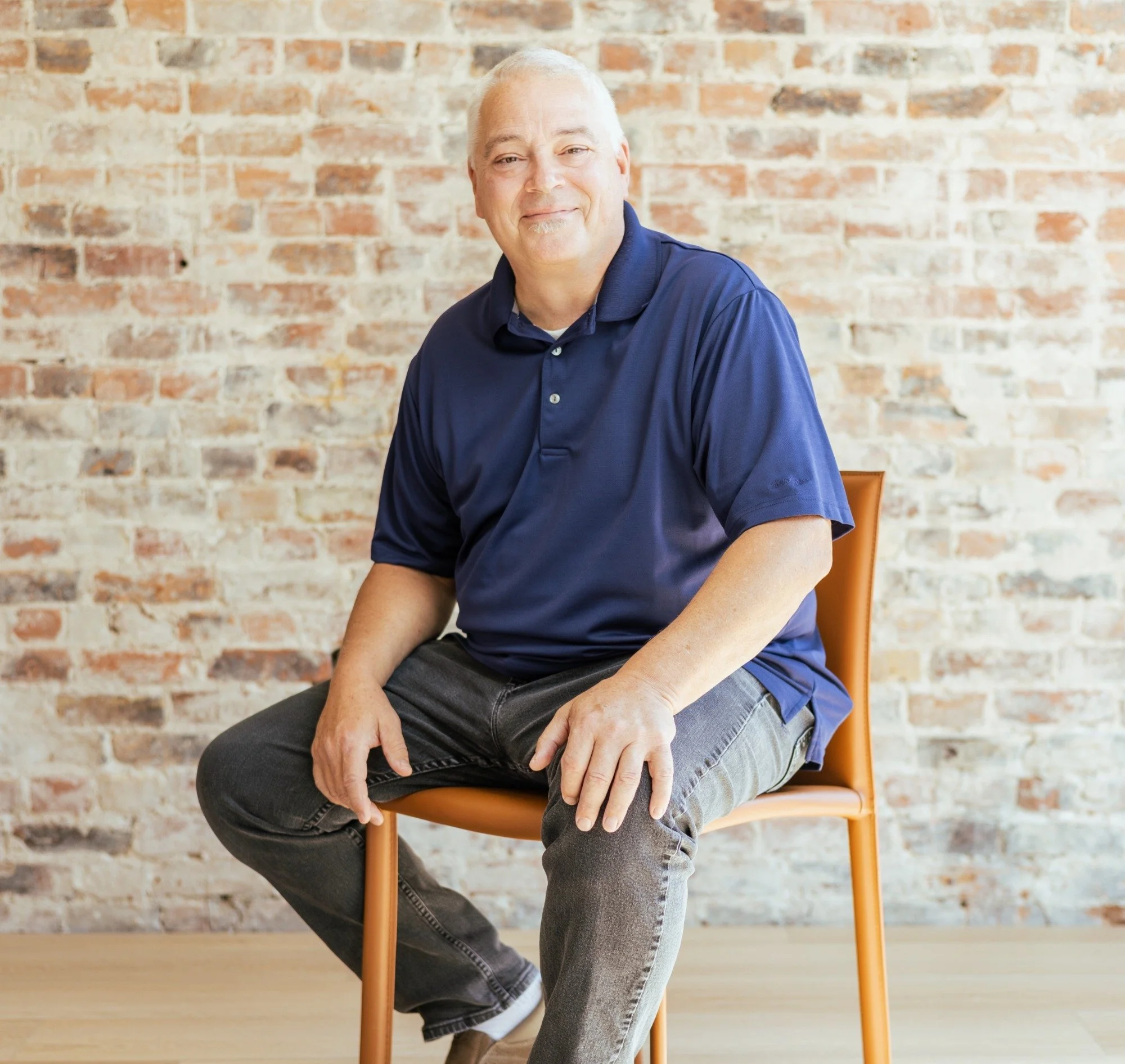 A middle-aged man sitting on an orange chair against a brick wall. He has short gray hair, a light smile, and is wearing a navy blue polo shirt, gray jeans, and brown shoes.