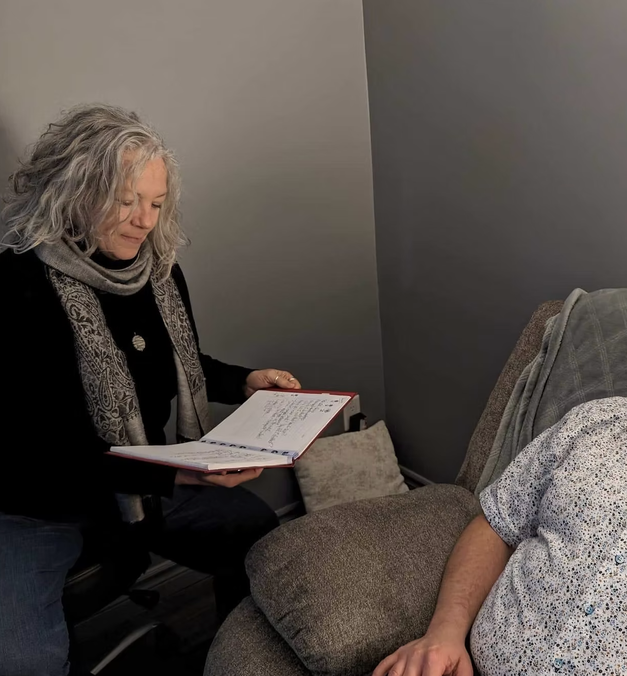 A woman with curly gray hair sitting on a chair, looking at an open notebook. Her arm is resting on an armrest of a patterned sofa where part of a person wearing a patterned shirt is visible.