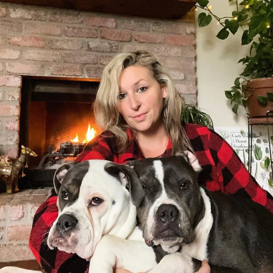 A woman with curly blonde hair sits in front of a fireplace with two Bully dogs, one white with black patches and the other black and white, in her lap.
