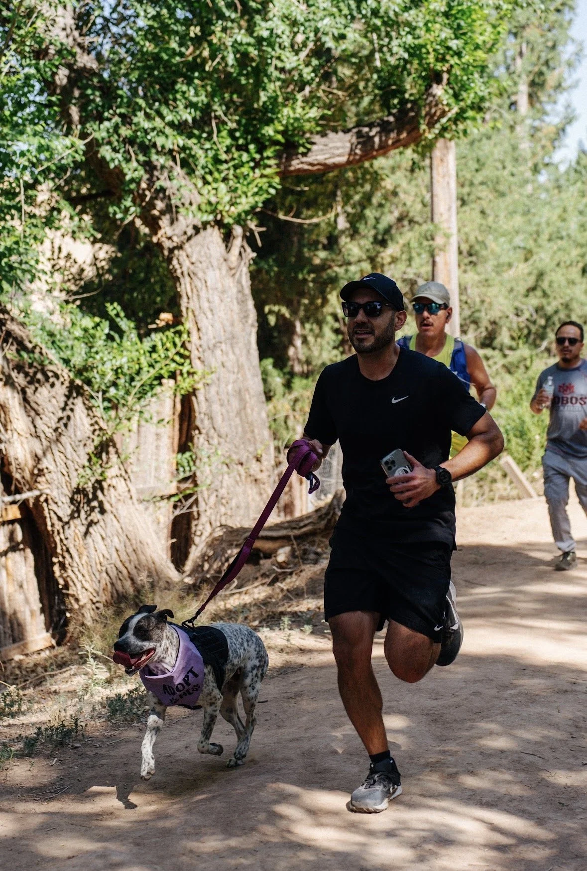 A man running on a dirt trail in a wooded area, holding a leash with a dog wearing a purple vest, with two other runners in the background.