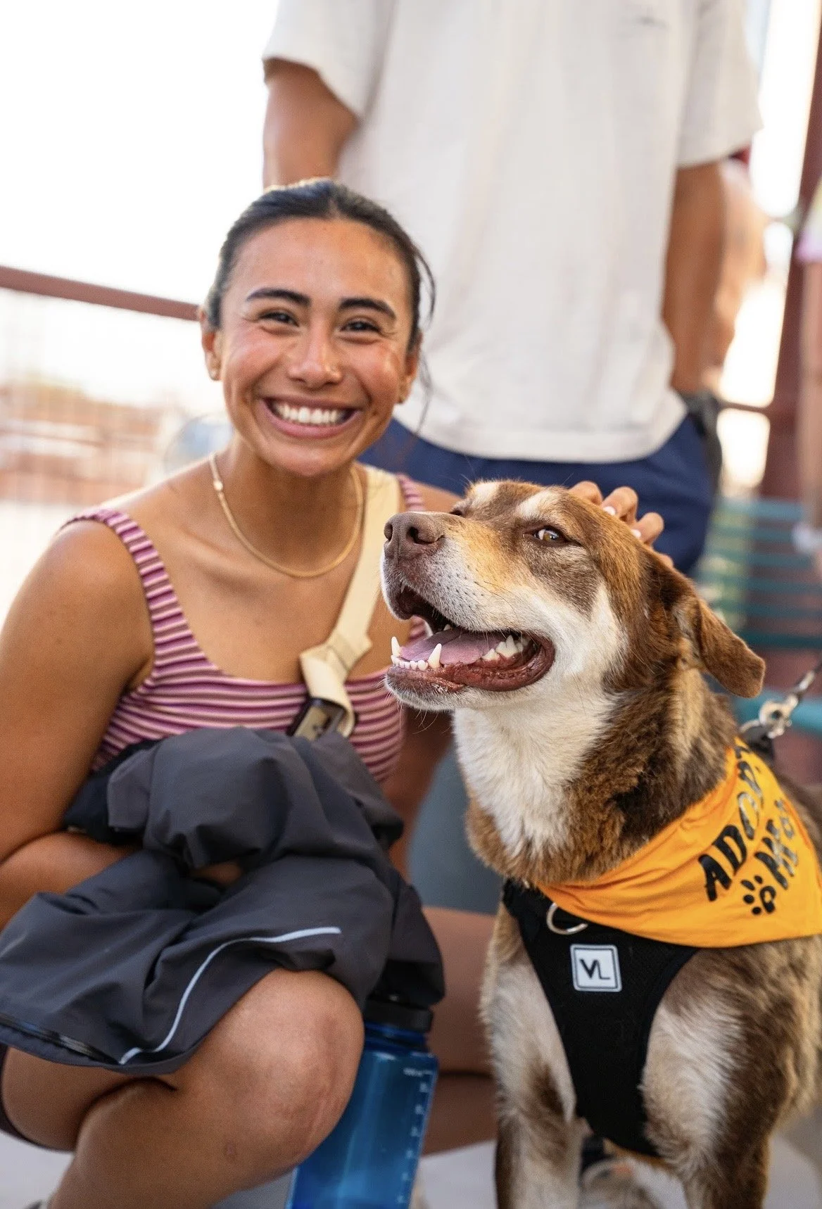 A woman smiling with a happy dog wearing an orange bandana, at an outdoor location.