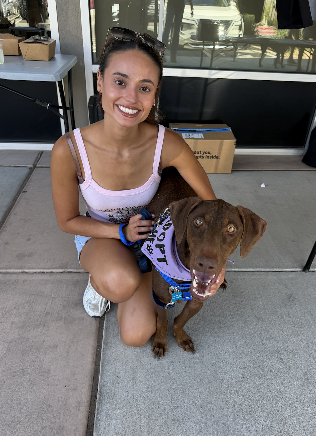 A smiling young woman kneeling on a sidewalk with a happy brown dog wearing a purple bandana that says "Service Dog". The woman has sunglasses on her head, a tank top, and shorts, and is holding the dog’s leash.