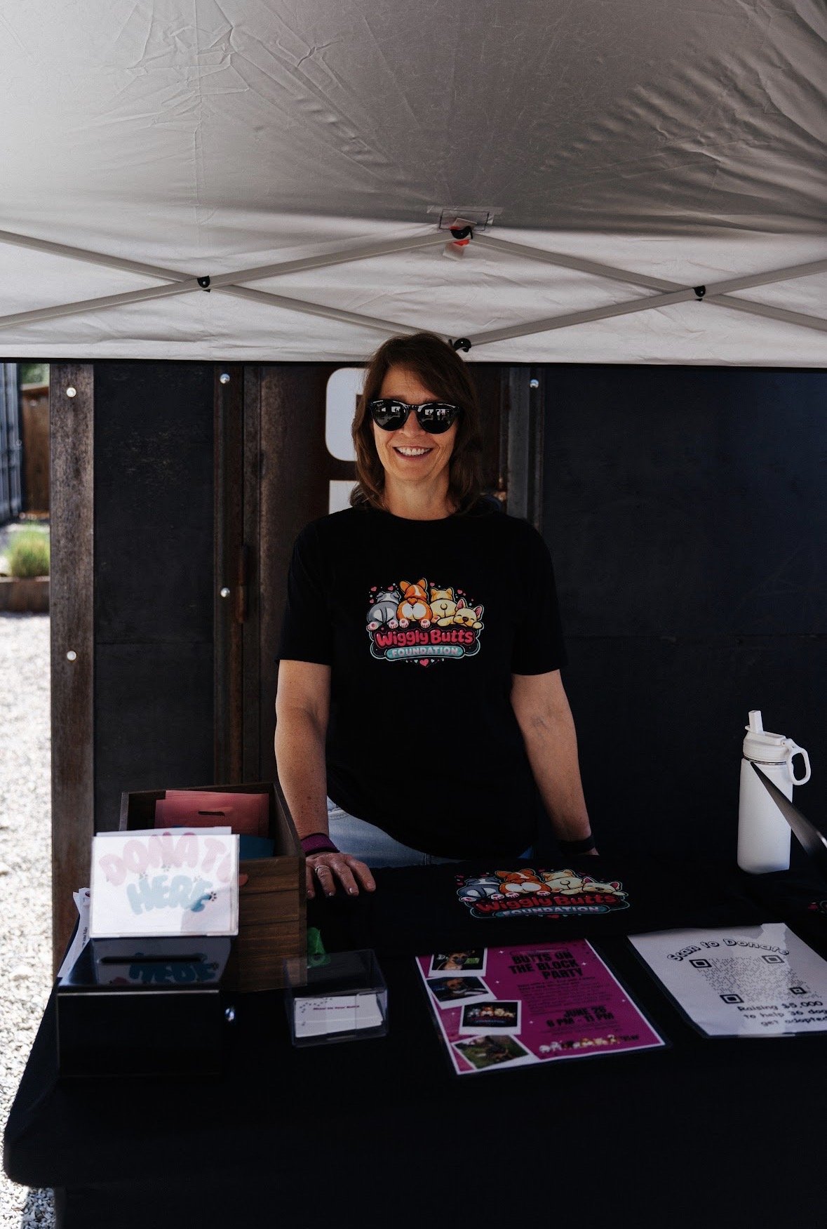 A woman is smiling and standing behind a table at an outdoor event. She is wearing sunglasses and a black t-shirt with cartoon dog characters and the text "Wiggly Butts Foundation." There are flyers, QR codes, and a water bottle on the table, which is under a white canopy.