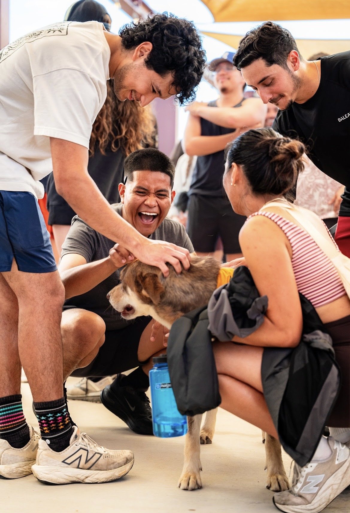 Group of people smiling and petting a dog indoors, with some standing and others squatting, wearing casual clothes and sneakers.
