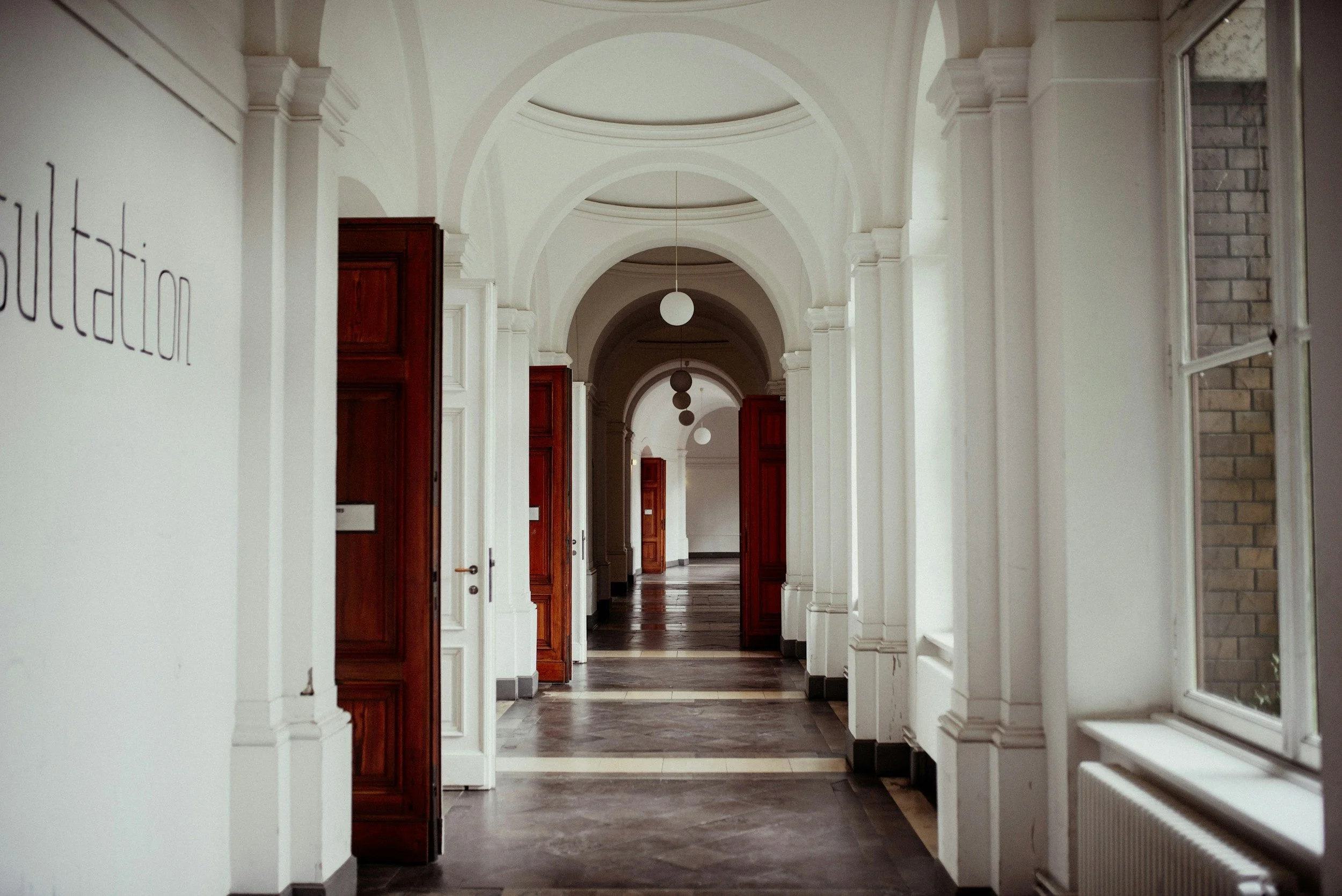A long hallway with white walls, arched ceiling, wooden doors, and hanging spherical light fixtures.