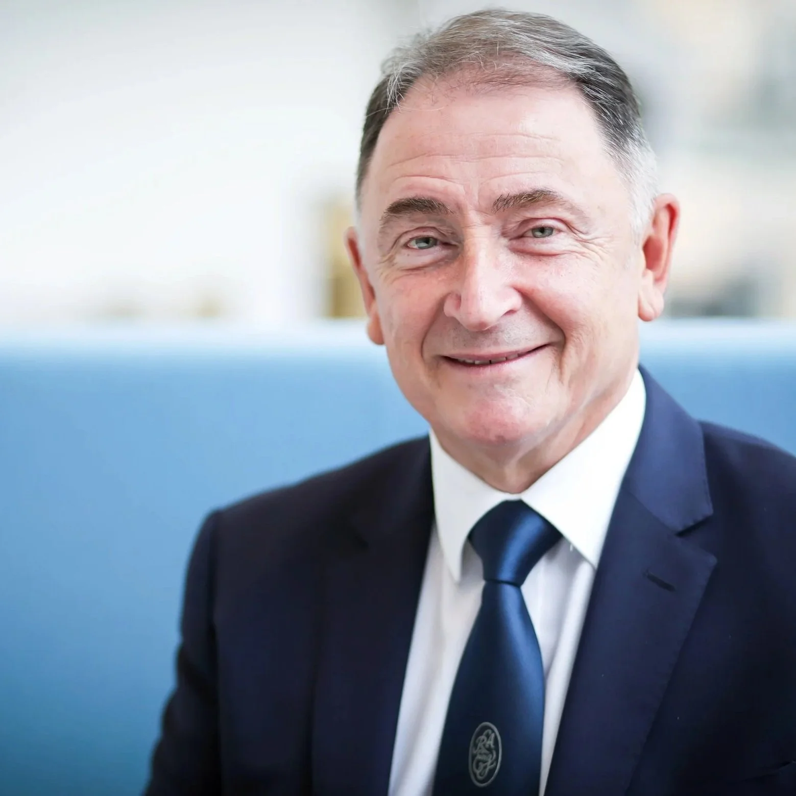 A middle-aged man with gray hair and blue eyes, dressed in a dark suit, white shirt, and blue tie, smiling in an indoor setting with a blurred background.