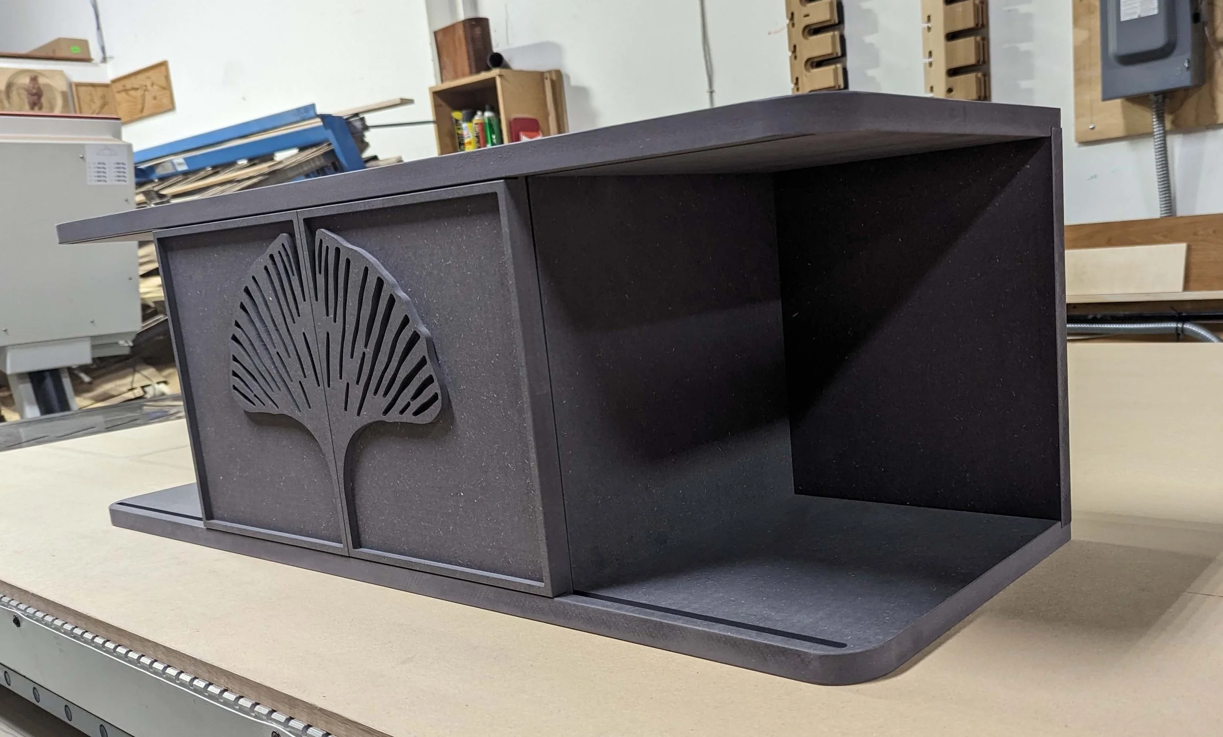 A black wooden storage box with a leaf-shaped cutout on the sliding doors, sitting on a workbench in a workshop.