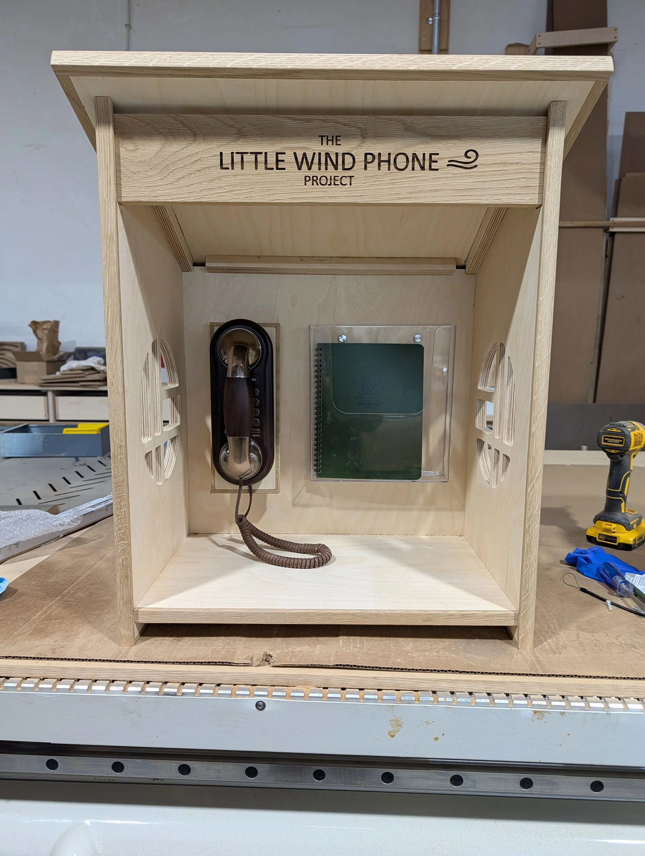 A wooden display case labeled 'The Little Wind Phone Project' housing an antique black rotary phone, a spiral notebook, and a transparent display stand, with workshop tools visible in the background.