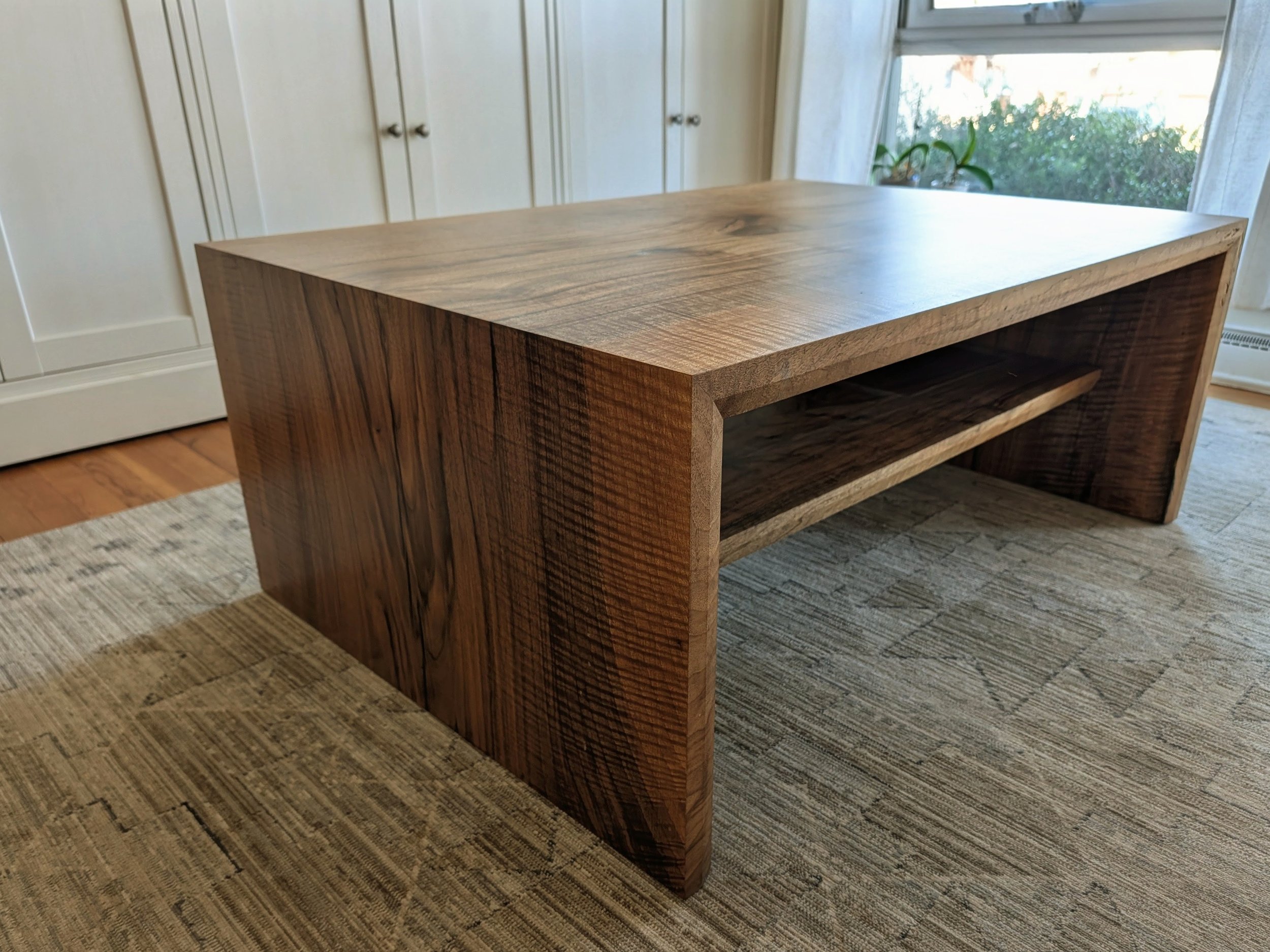 A wooden coffee table with a thick tabletop and a lower shelf, situated on a beige rug in a room with white cabinetry and a large window with a view of greenery outside.