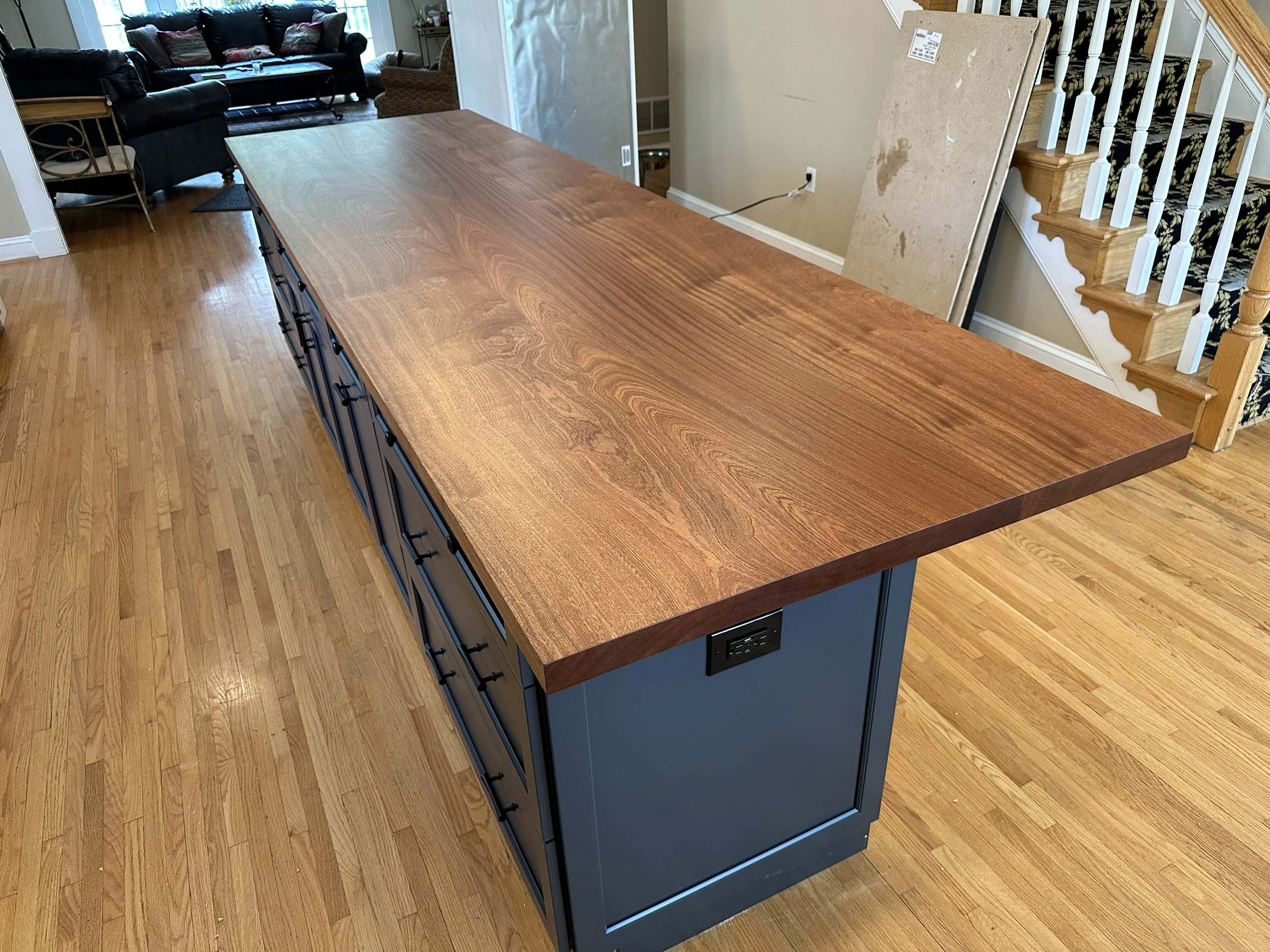 A large kitchen island with a wooden countertop and blue cabinet base, located in a home with hardwood floors, adjacent to a staircase with white spindles and a black carpet runner.