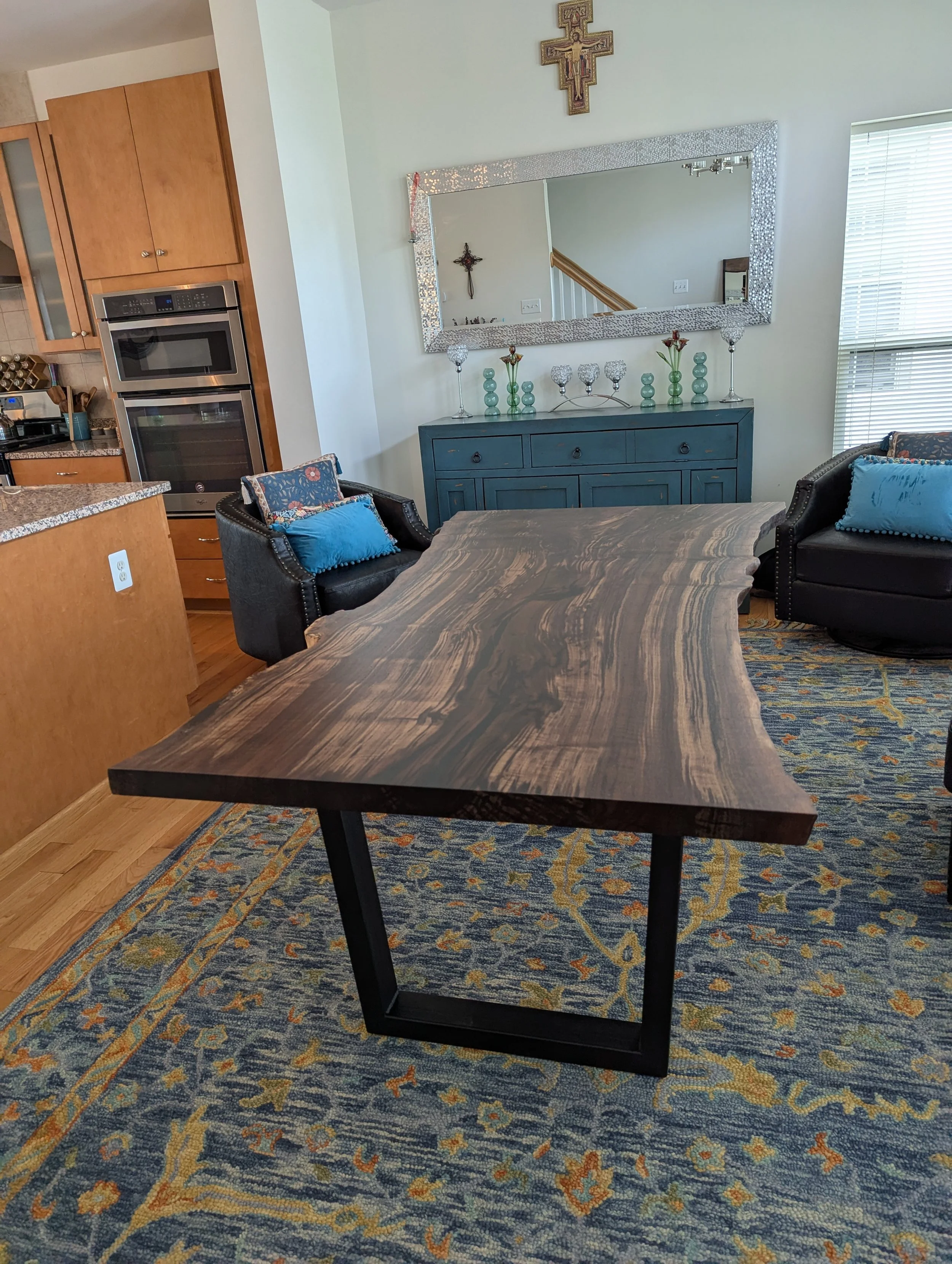 Living room with a wooden table, black leather armchairs with blue pillows, a blue sideboard with decorative glass items, a large mirror, religious crosses on the wall, and a kitchen area with wooden cabinets in the background.
