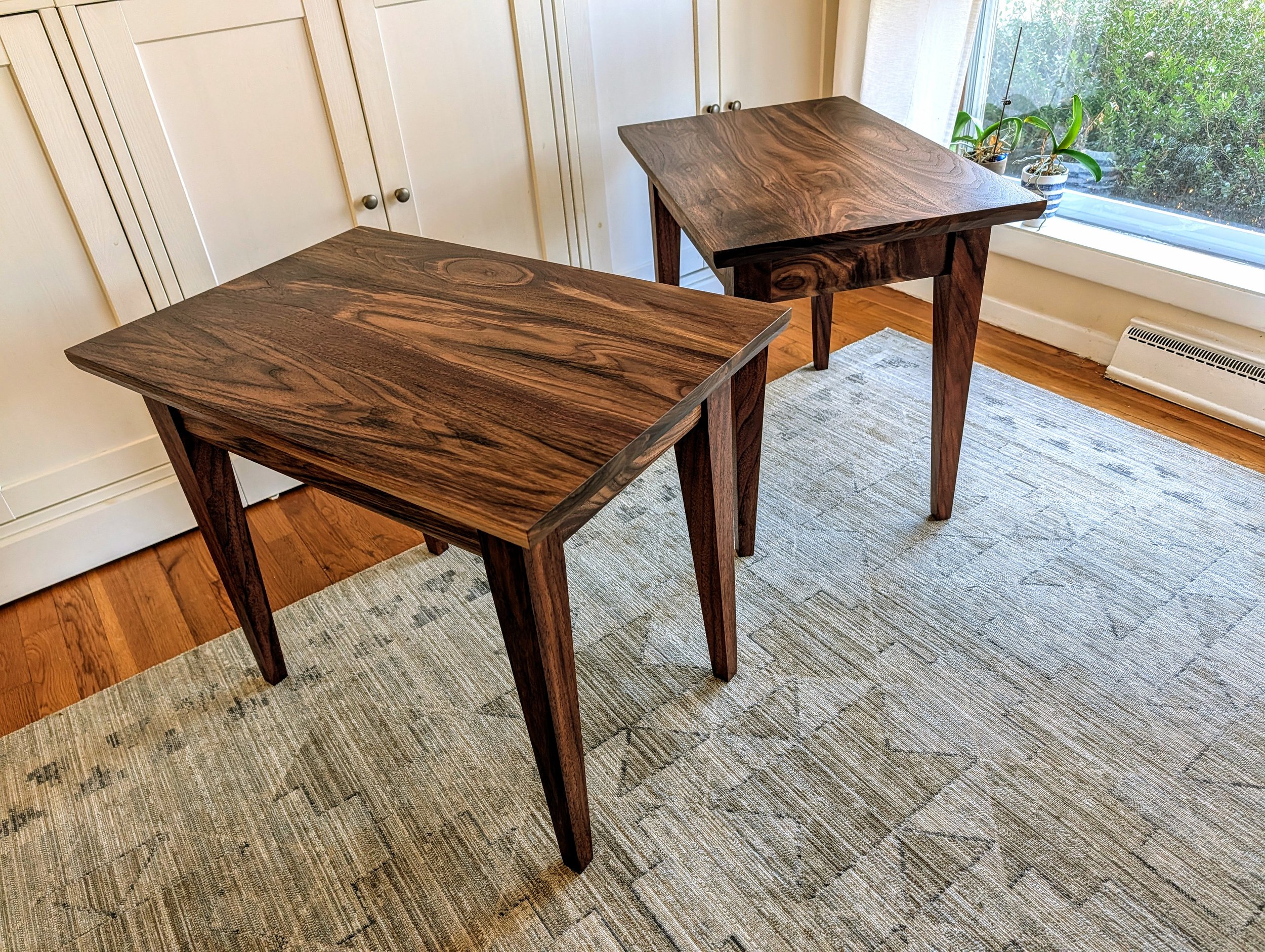 Two wooden tables with dark wood grain pattern in a room with white cabinets and a window with plants outside.