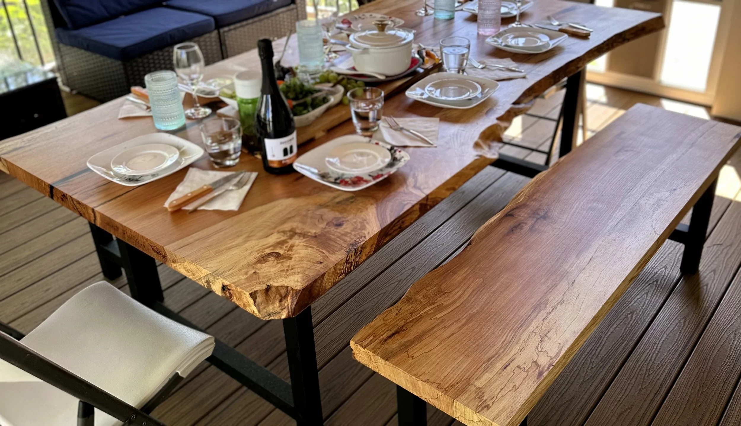 A wooden dining table set for a meal with plates, glasses, a bottle of wine, and decorative items, on a deck with outdoor chairs.
