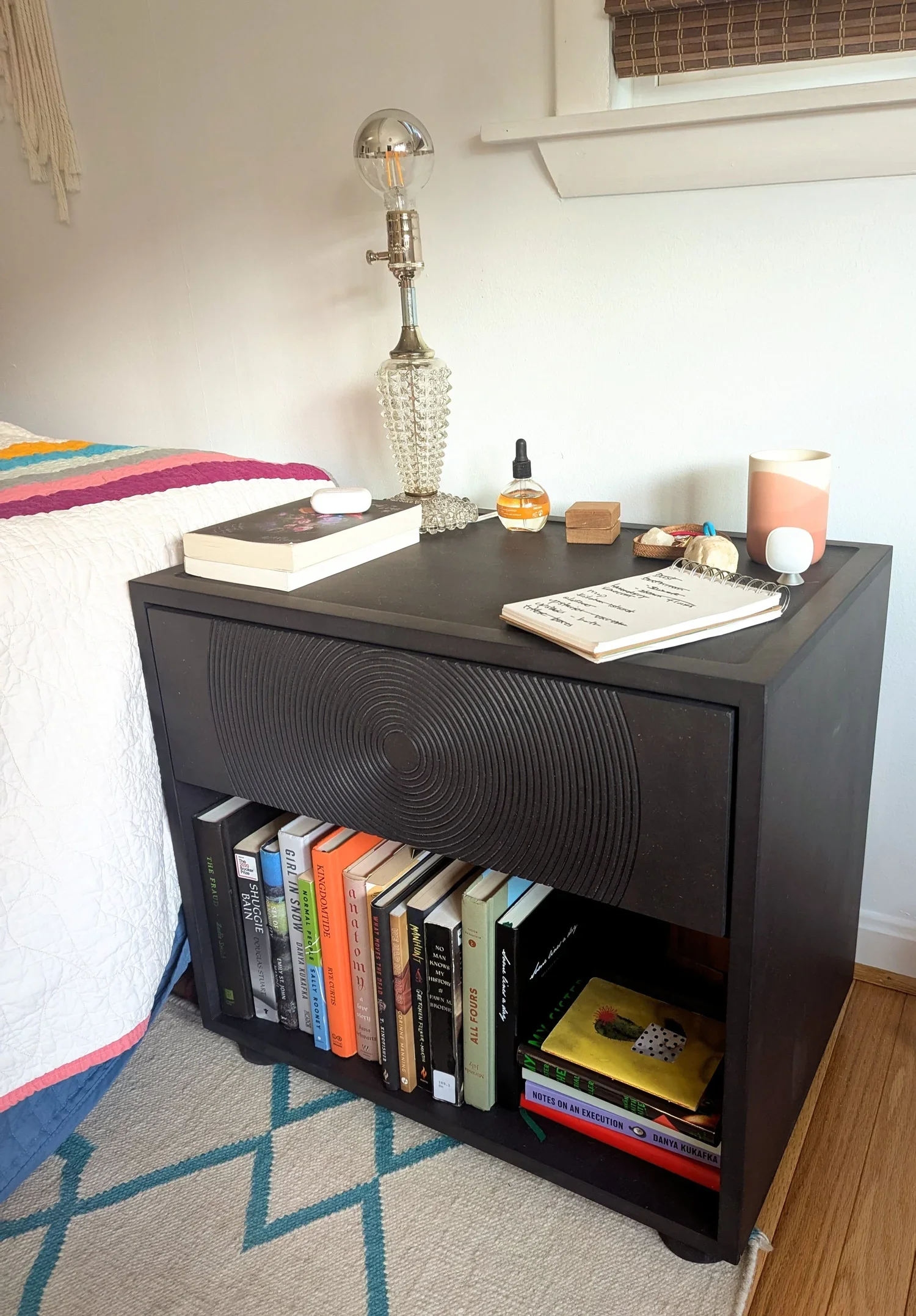 Black bookshelf bedside table with books, decorative lamp, notebook, and other small items, next to a bed with a colorful quilt, on a patterned rug.