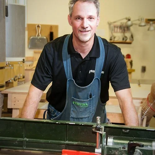 Chris Johnstone wears a black shirt and blue apron and smiles in a woodworking workshop, surrounded by tools and wooden projects.