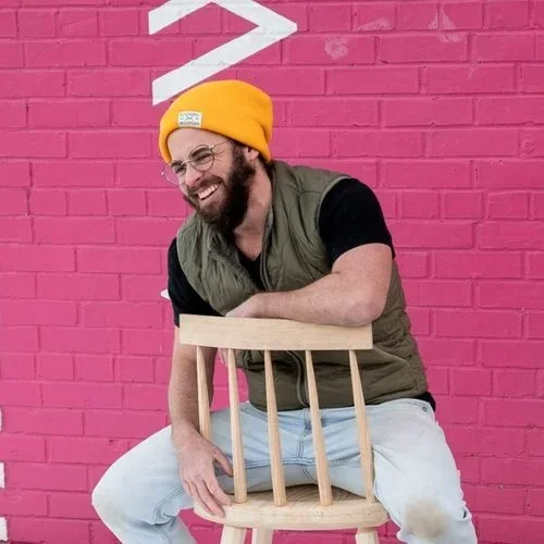 A man with a beard, glasses, and a yellow beanie, smiling and sitting on a wooden chair in front of a pink brick wall.