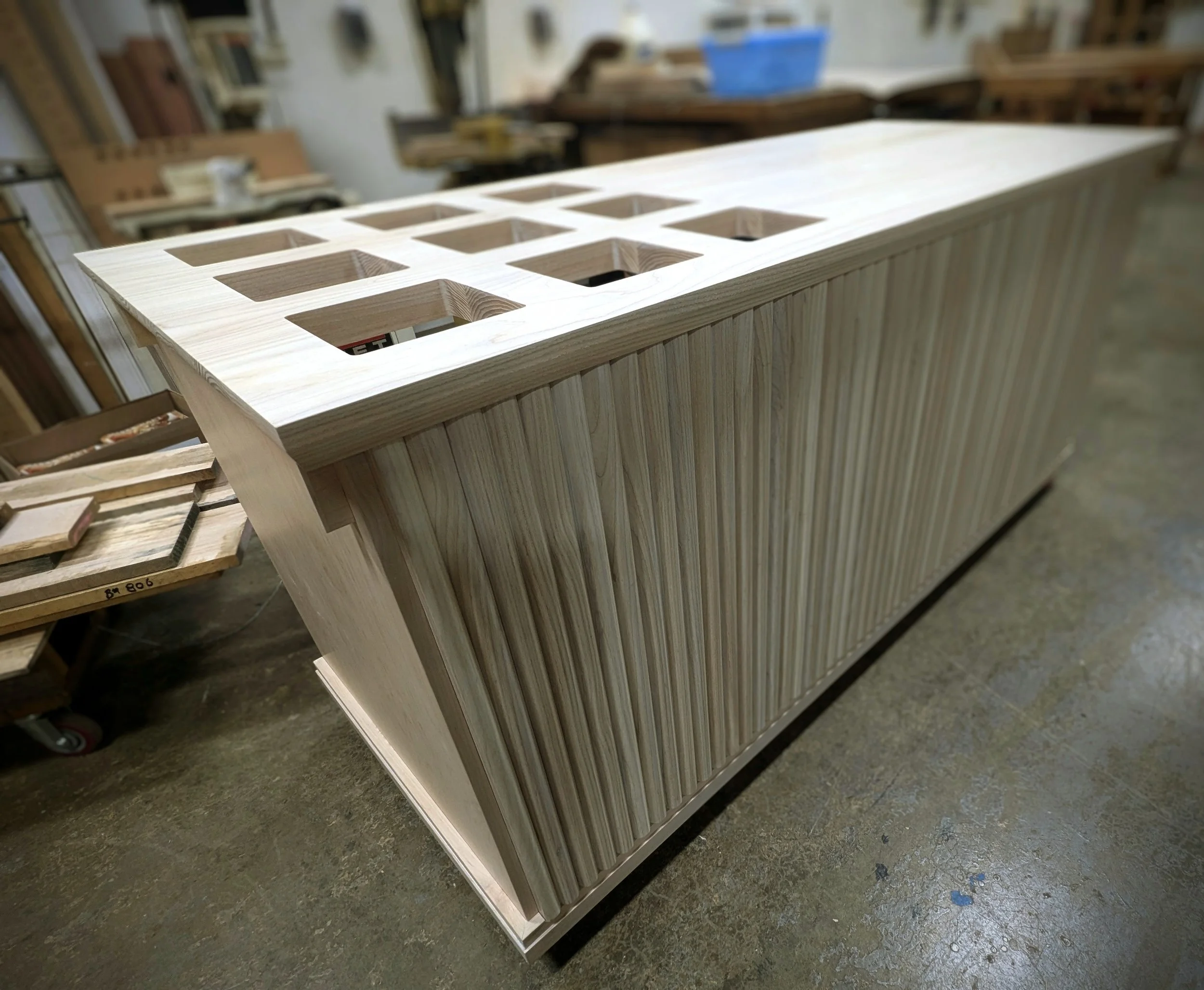 A woodworking project in progress, featuring a wooden structure with a top panel that has square cutouts, possibly for wine or bottle storage, in a workshop setting.