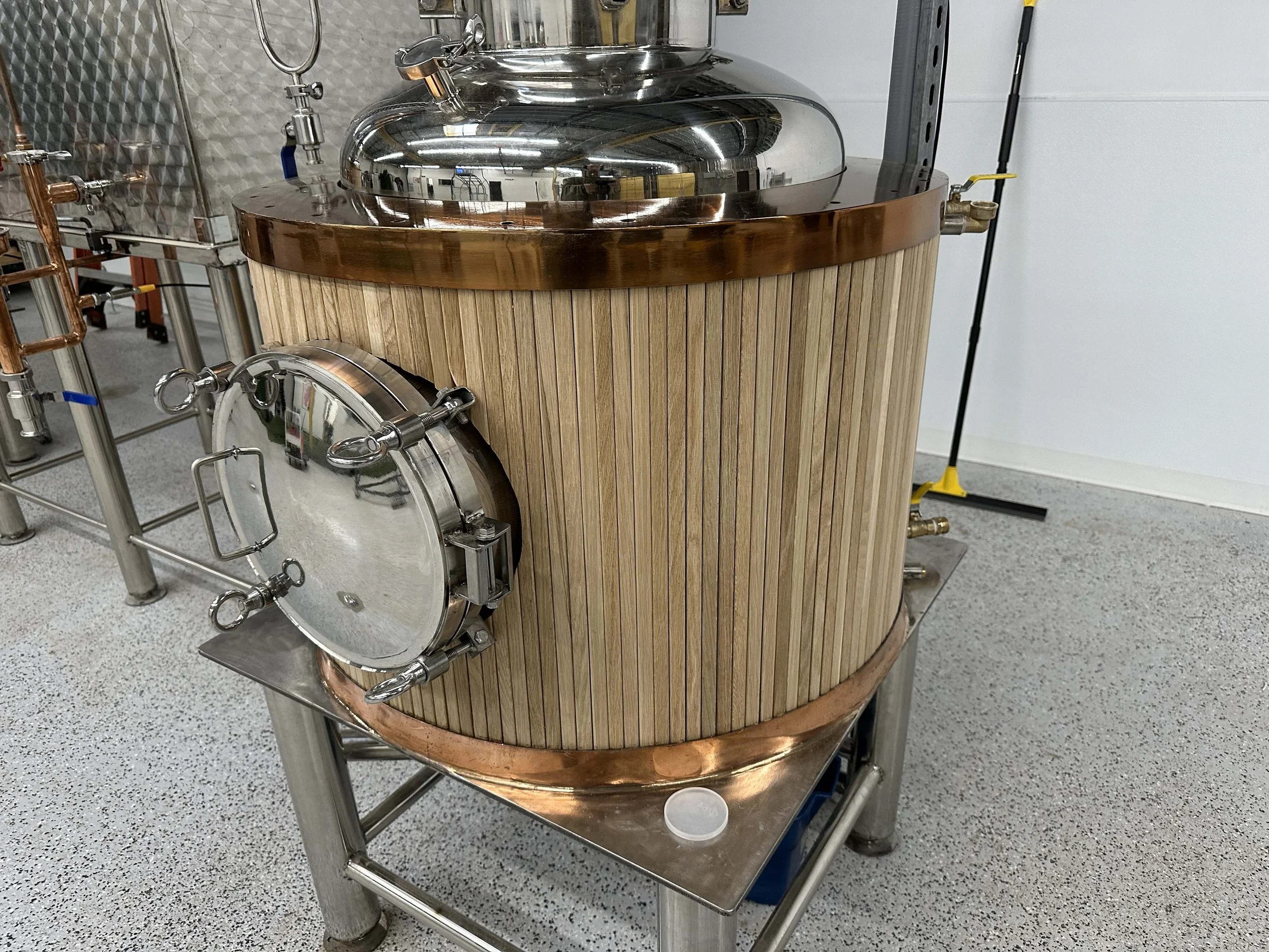 Copper and steel brewing vessel with a wooden exterior, situated in a brewery or distillery, with a door at the front for access.