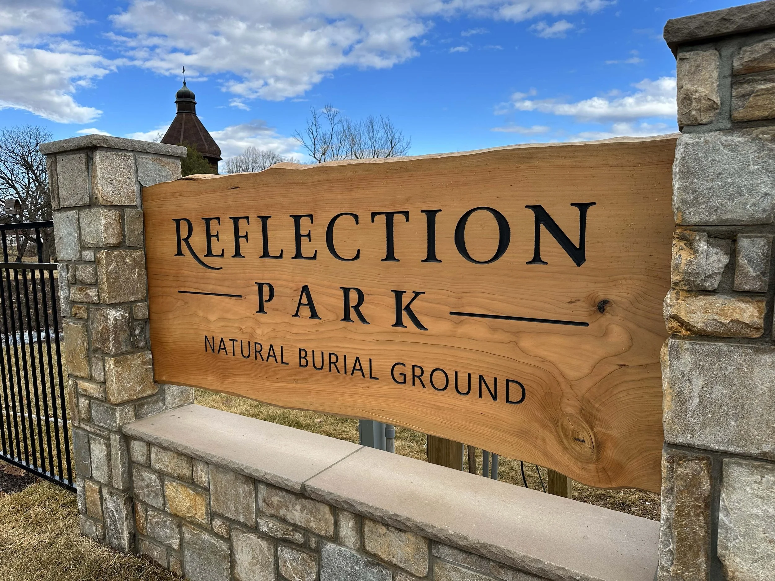 Wooden sign constructed from an 8 foot long, live edge cedar slab with black lettering, mounted on stone pillars, with a fence and a building with a tower in the background, under a partly cloudy blue sky.