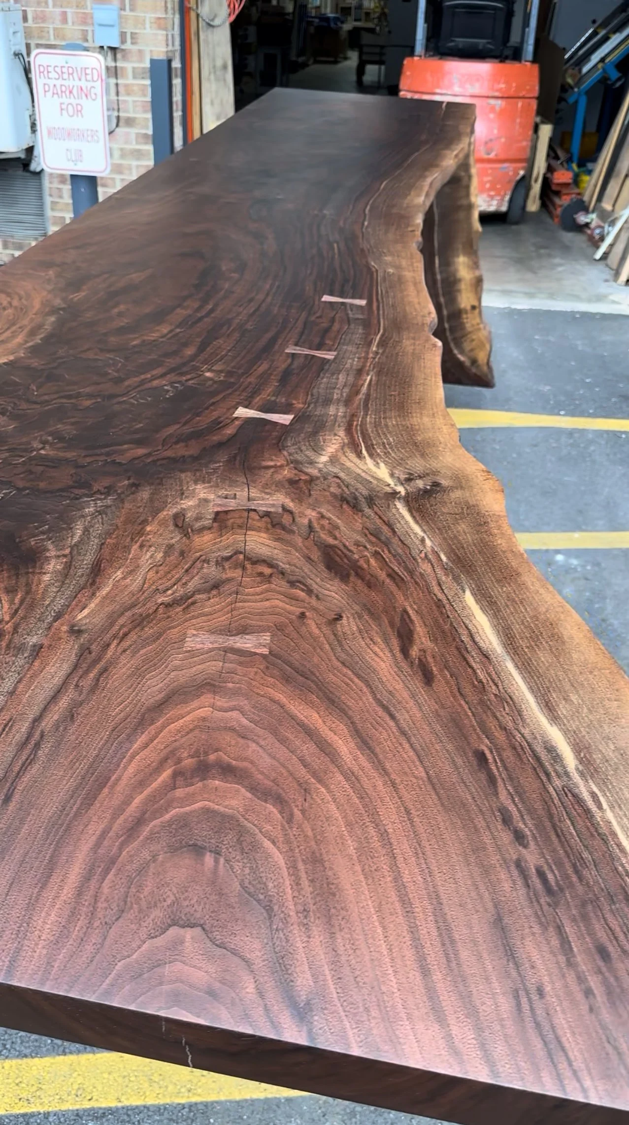 A large, polished wooden slab with a natural, irregular edge, placed outdoors near a workshop or hardware store.