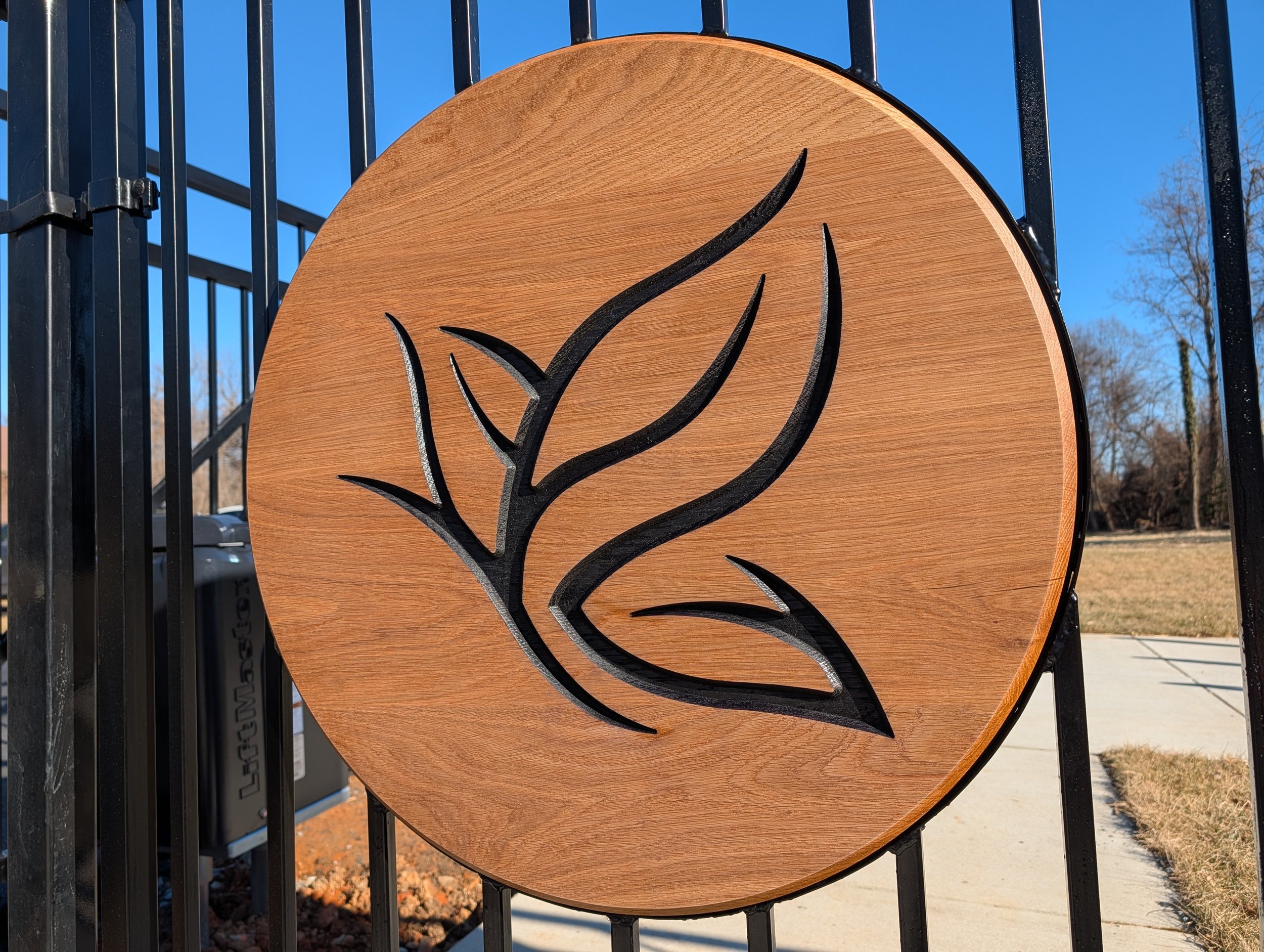Wooden sign with a black engraved leaf design, mounted on a black metal fence outside.