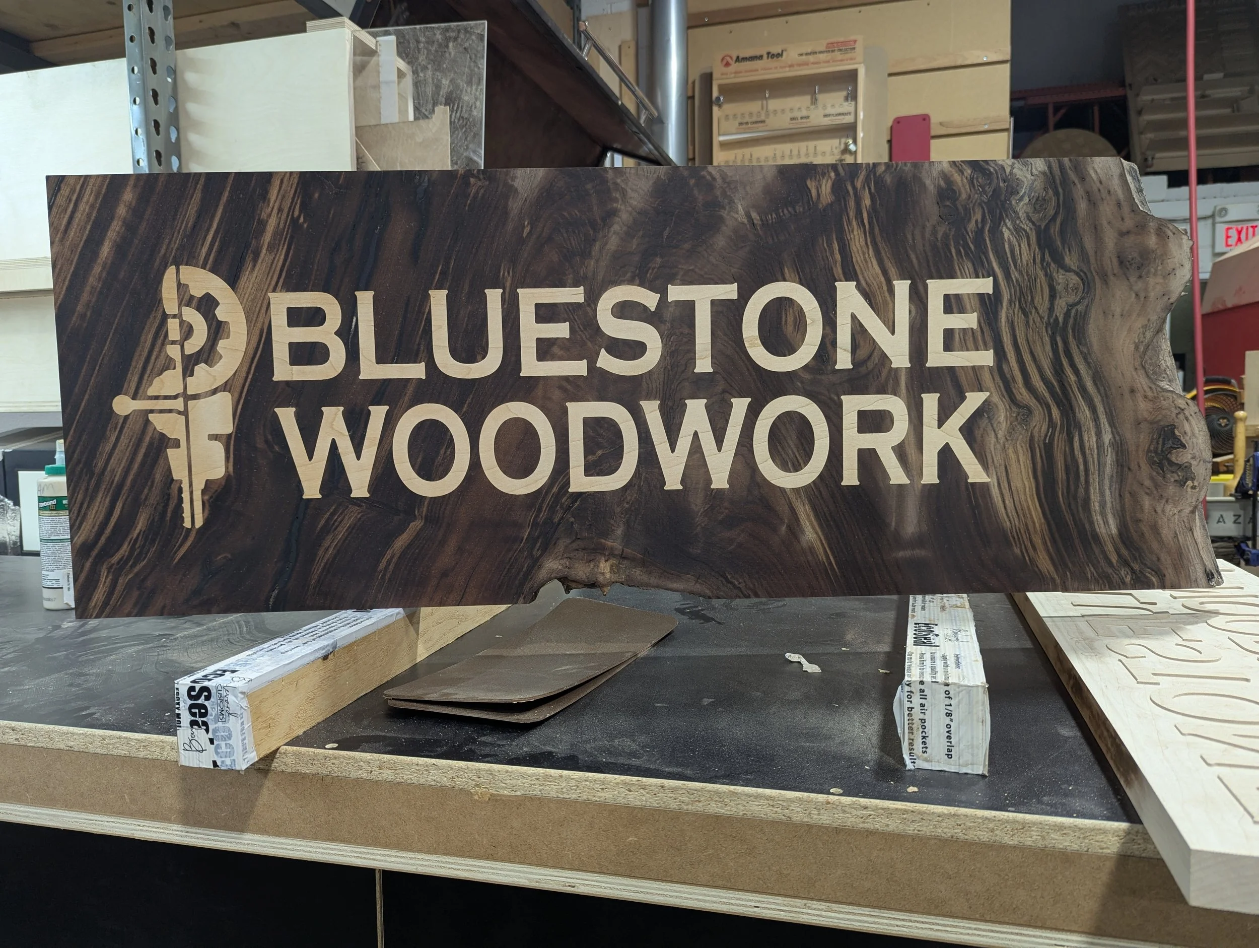 A woodworking sign that reads 'BLUESTONE WOODWORK' with a logo of a worker cutting a piece of wood, carved into a dark, natural wood slab.
