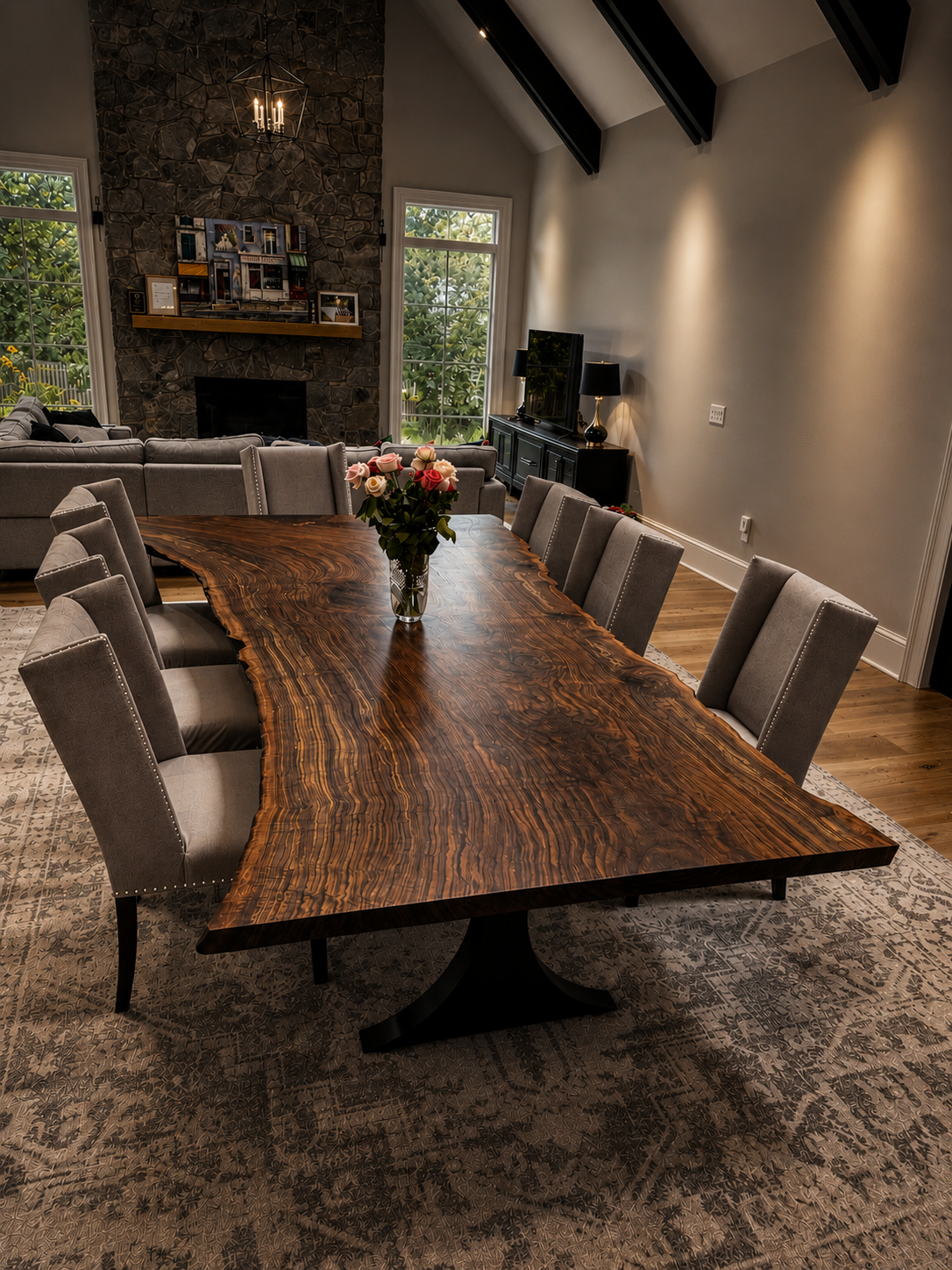 12-foot live-edge claro walnut dining table with a black pedestal base, seating ten gray upholstered chairs, in a great room with a stone fireplace