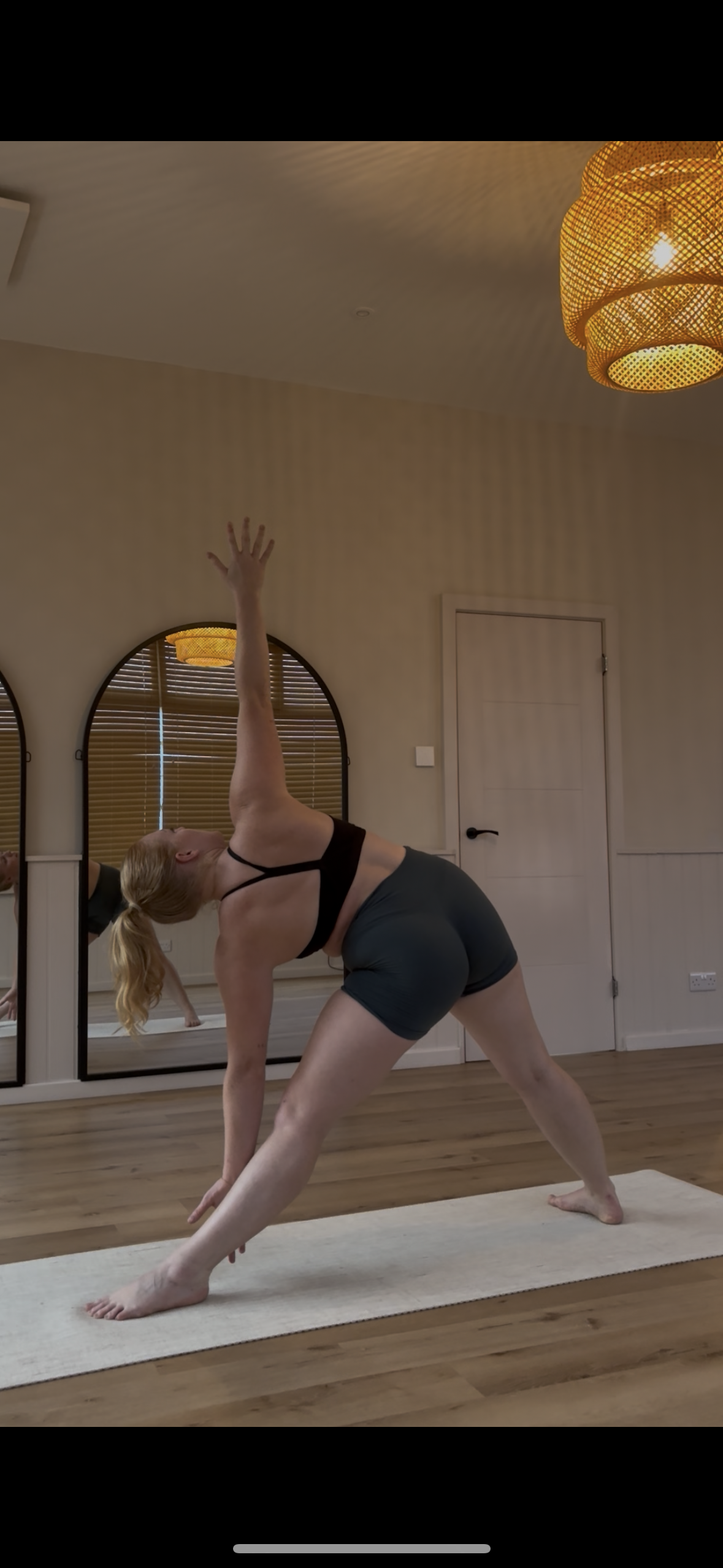 A woman practicing yoga indoors, performing a side angle pose on a white yoga mat, with a mirror and wooden blinds behind her, in a room with wooden flooring and warm lighting.