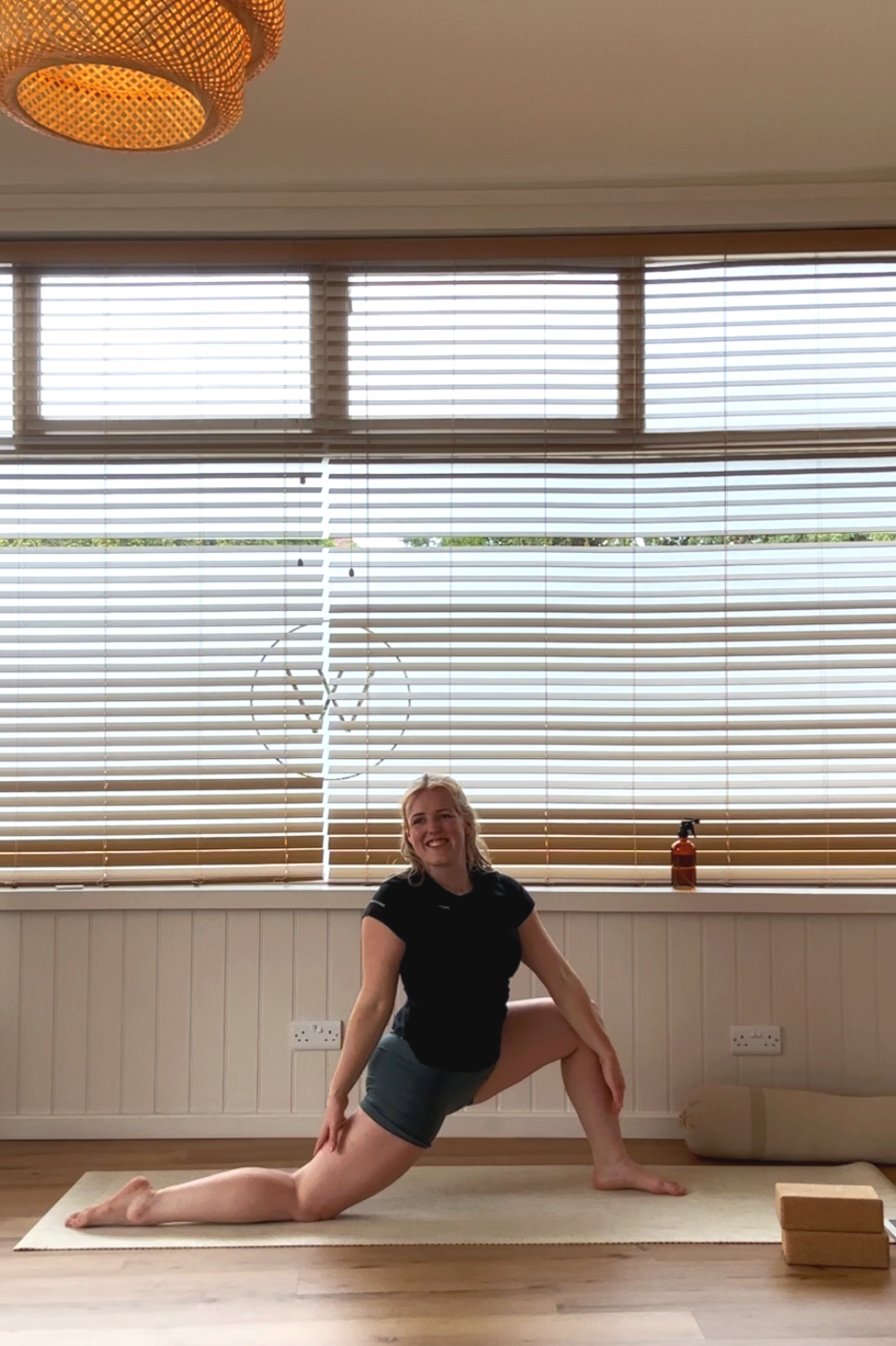 A woman practicing yoga indoors on a cream-colored mat, wearing a black top and gray shorts, smiling and in a lunge pose with one knee on the ground and both hands resting on her bent knee, in front of a window with horizontal blinds.