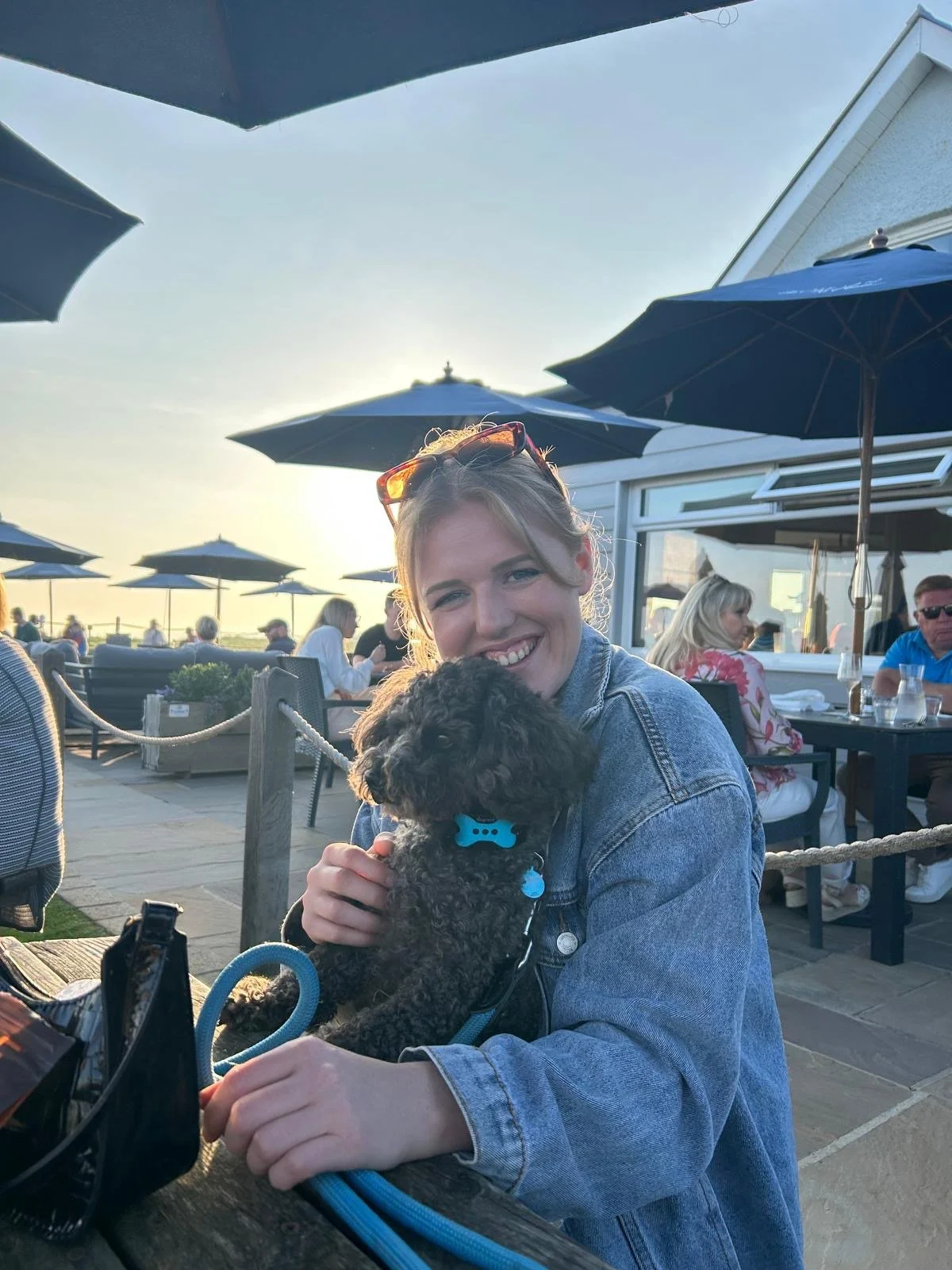 A woman in a denim jacket smiling and holding a small black curly-haired dog with a blue collar, seated at an outdoor restaurant patio during sunset with umbrellas and other diners in the background.
