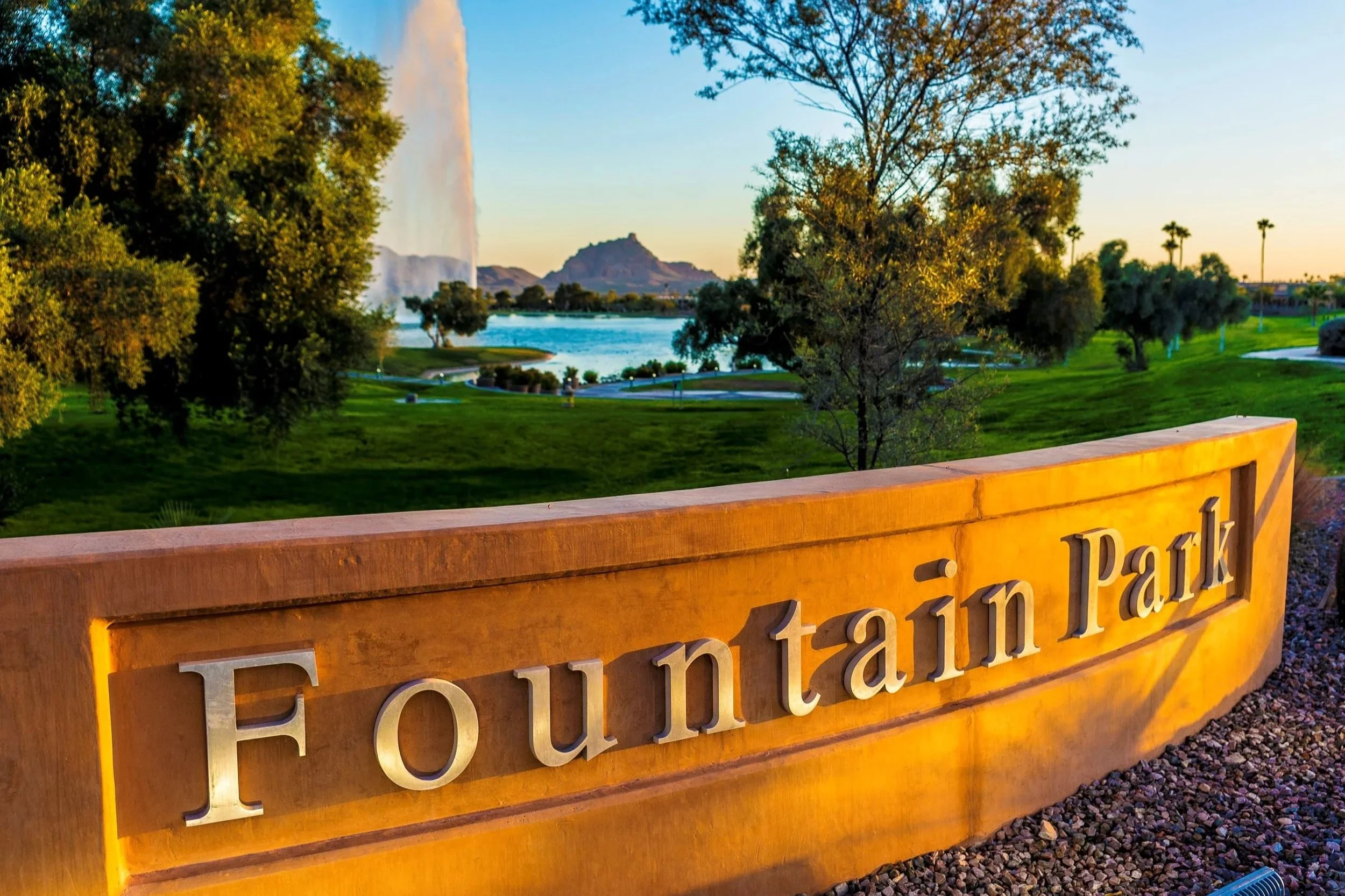 A large stone sign reads 'Fountain Park' in front of a lush green park with trees, a lake, and a fountain. In the background, mountains and a partly cloudy sky are visible during sunset.