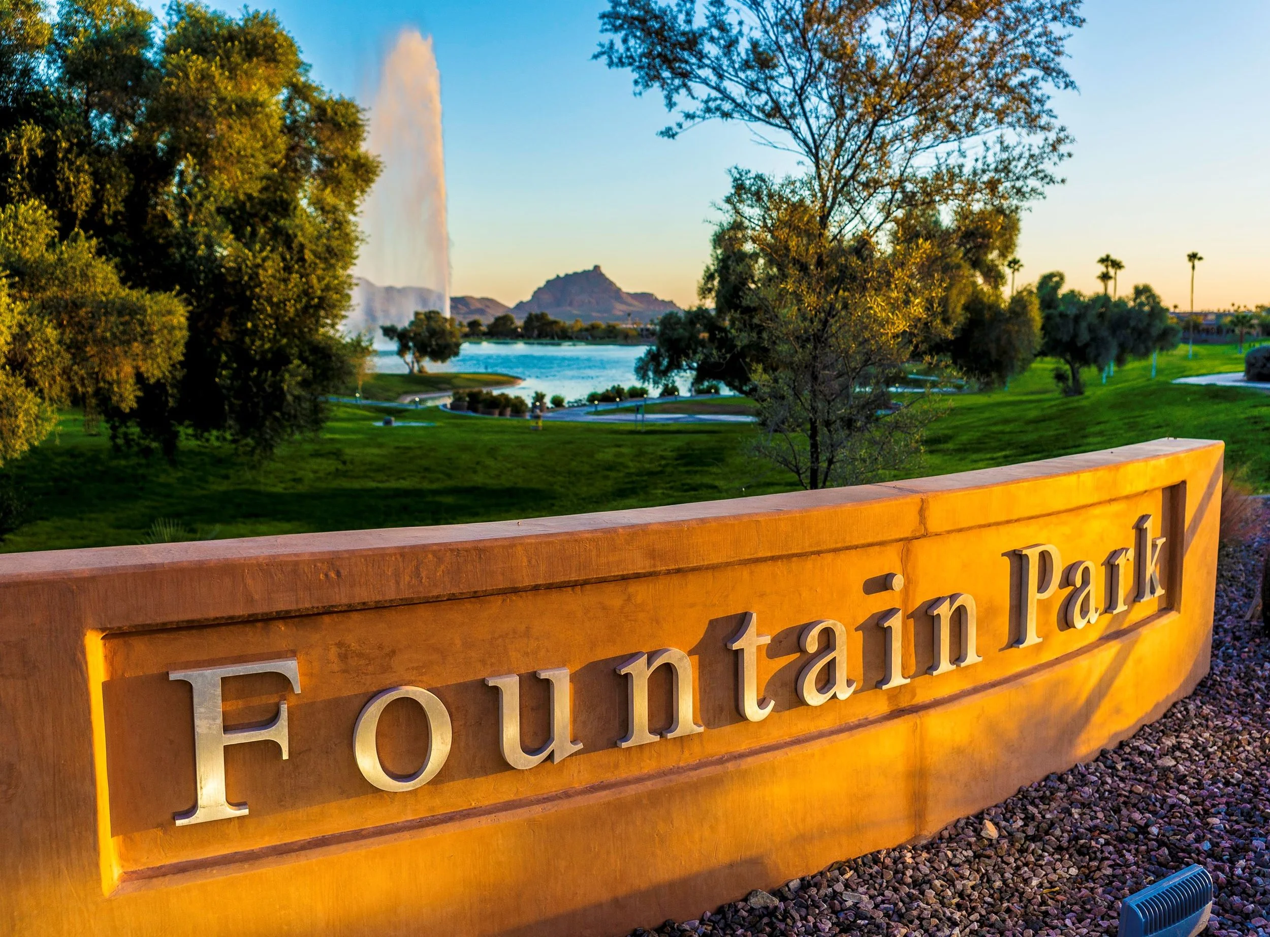 A large stone sign reading 'Fountain Park' with metal letters, situated in a park at sunset. In the background, there is a pond with a fountain, lush green grass, trees, and mountains in the distance.