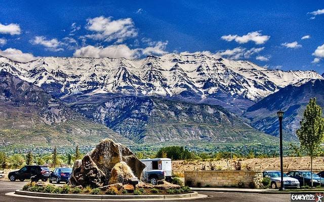 Mountain range with snow-capped peaks under a partly cloudy sky, in front a parking lot with cars, a large rock, and a few trees.