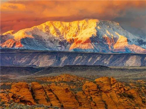 Snow-capped mountain illuminated by the sunset with rocky desert terrain in the foreground.