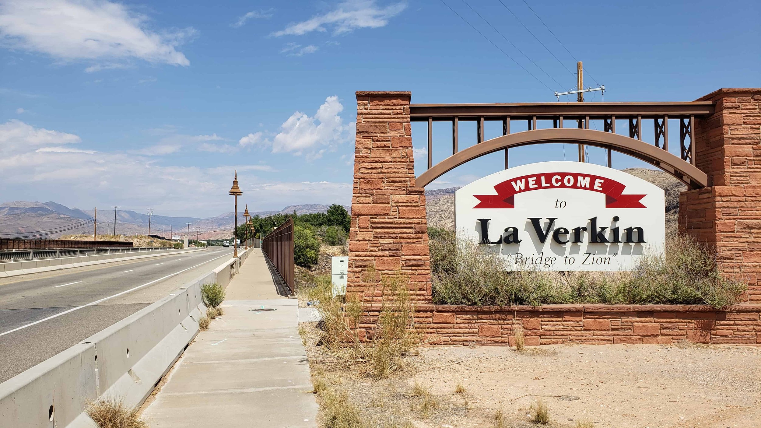 A welcome sign for La Verkin, Utah, with a red brick wall, a sidewalk, and a road with mountains and a blue sky in the background.