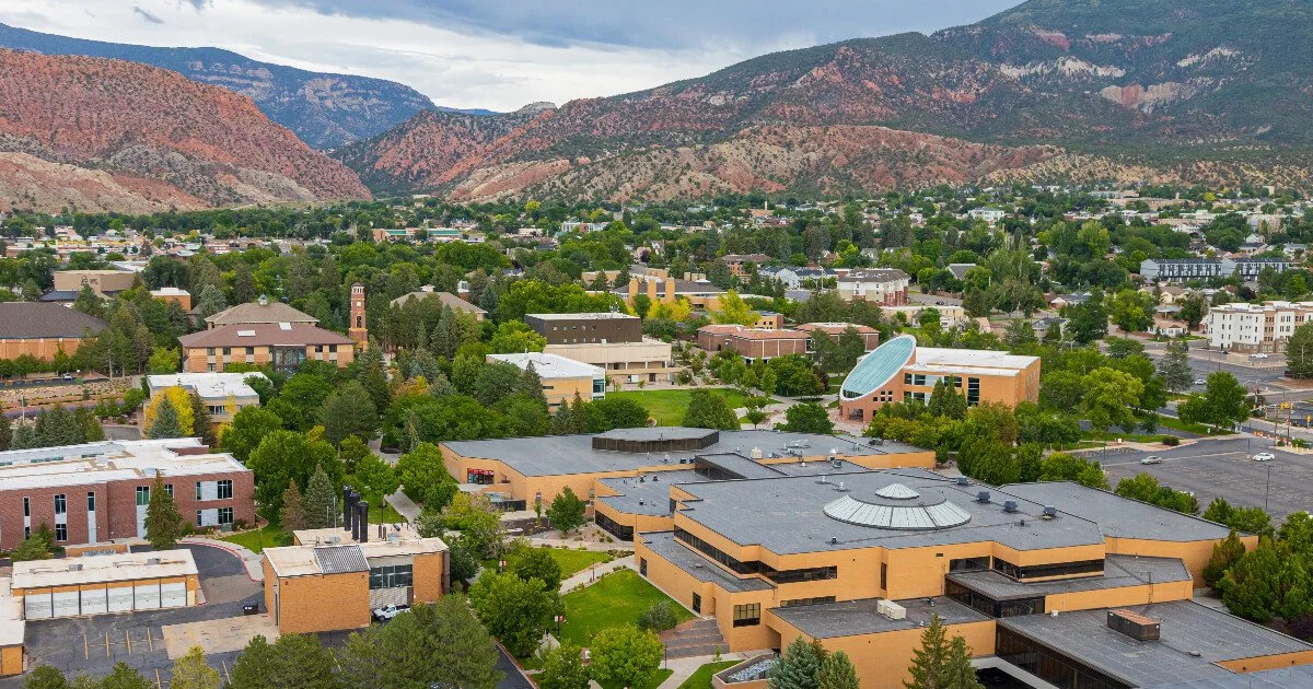 Aerial view of a city nestled in a valley with mountains in the background. The city features various buildings, trees, and parking lots.