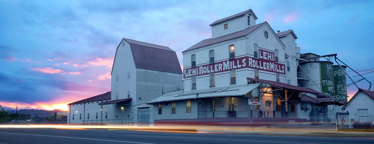 An old white barn with a large sign that reads 'Lehi Rollermills' at sunset, with streaks of light from passing cars in the foreground.
