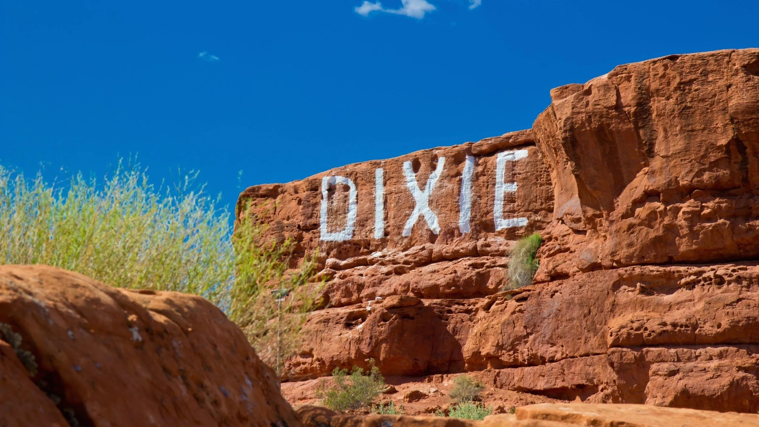 A red rock formation with the word "DIXIE" painted in white on it, against a bright blue sky with a few clouds, with some sparse green vegetation at the base.