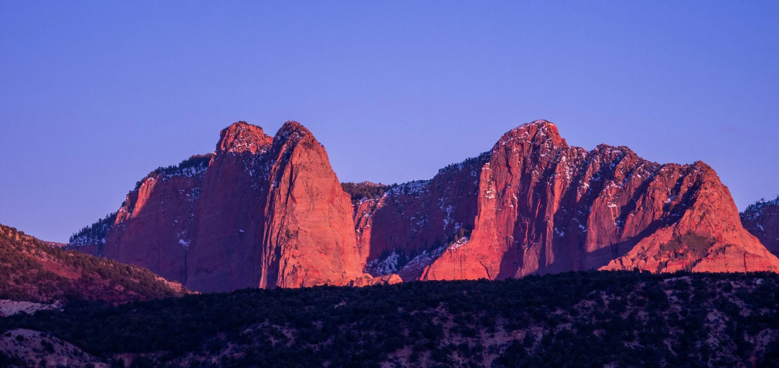 Sunset over red rock mountains with patches of snow and a clear blue sky.