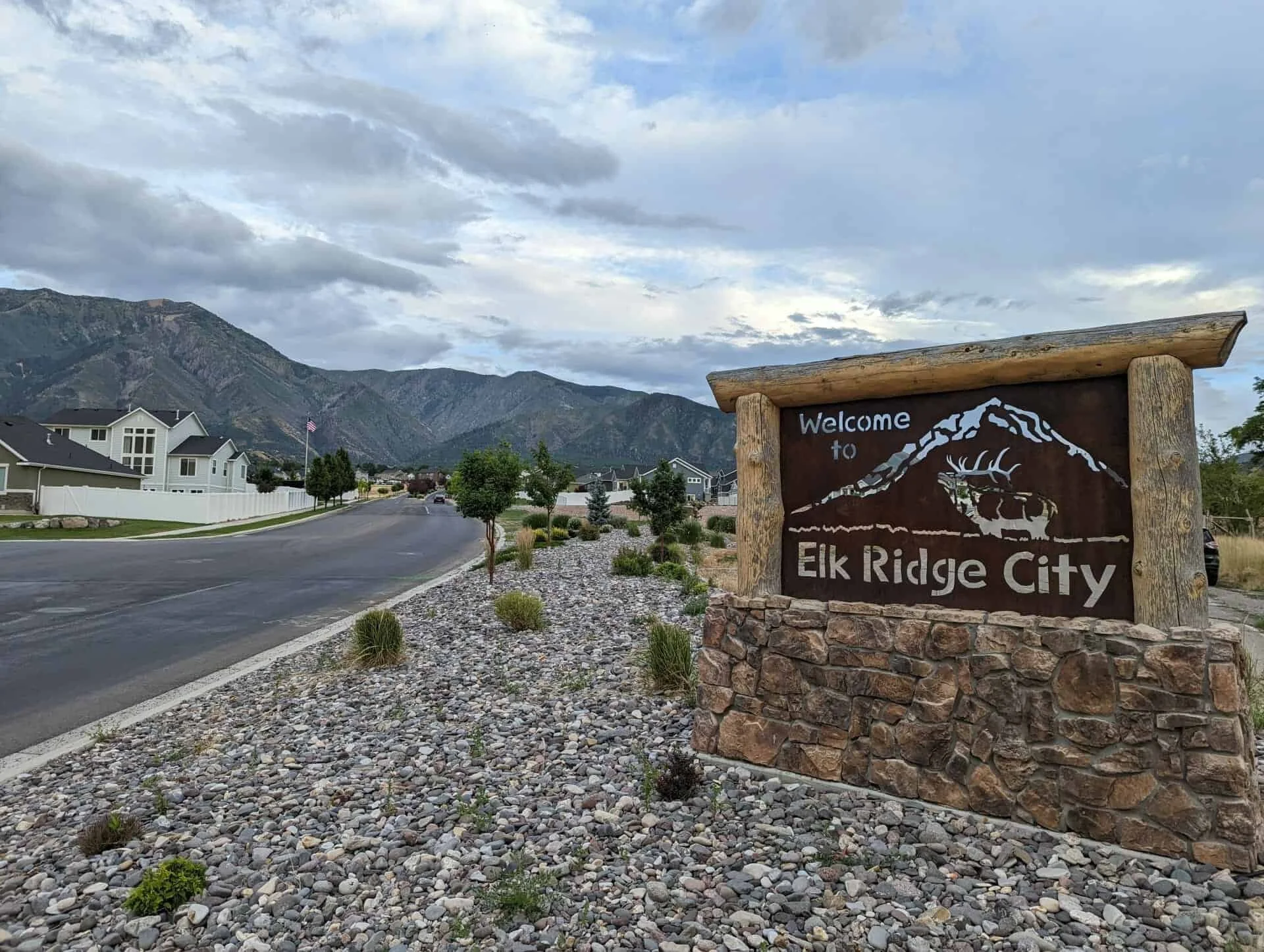 Welcome sign for Elk Ridge City with a mountain range and residential neighborhood in the background, cloudy sky, and landscaped rocks and small plants in the foreground.