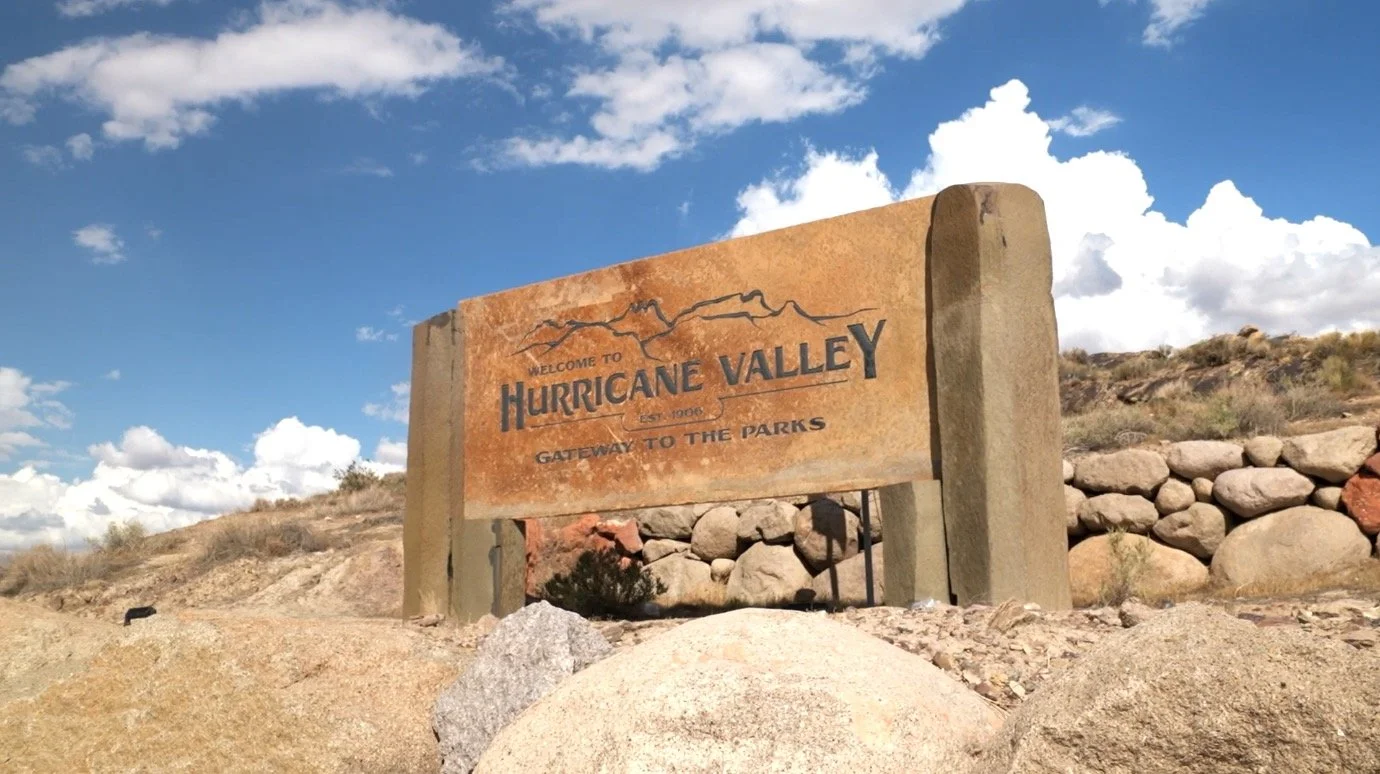 A welcome sign for Hurricane Valley that reads 'Gateway to the Parks,' outdoors with a blue sky, clouds, rocks, and dry desert vegetation in the background.