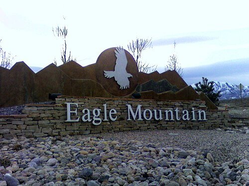 Entrance sign for Eagle Mountain with a white eagle silhouette against a mountain-shaped backdrop, desert landscape with rocks.