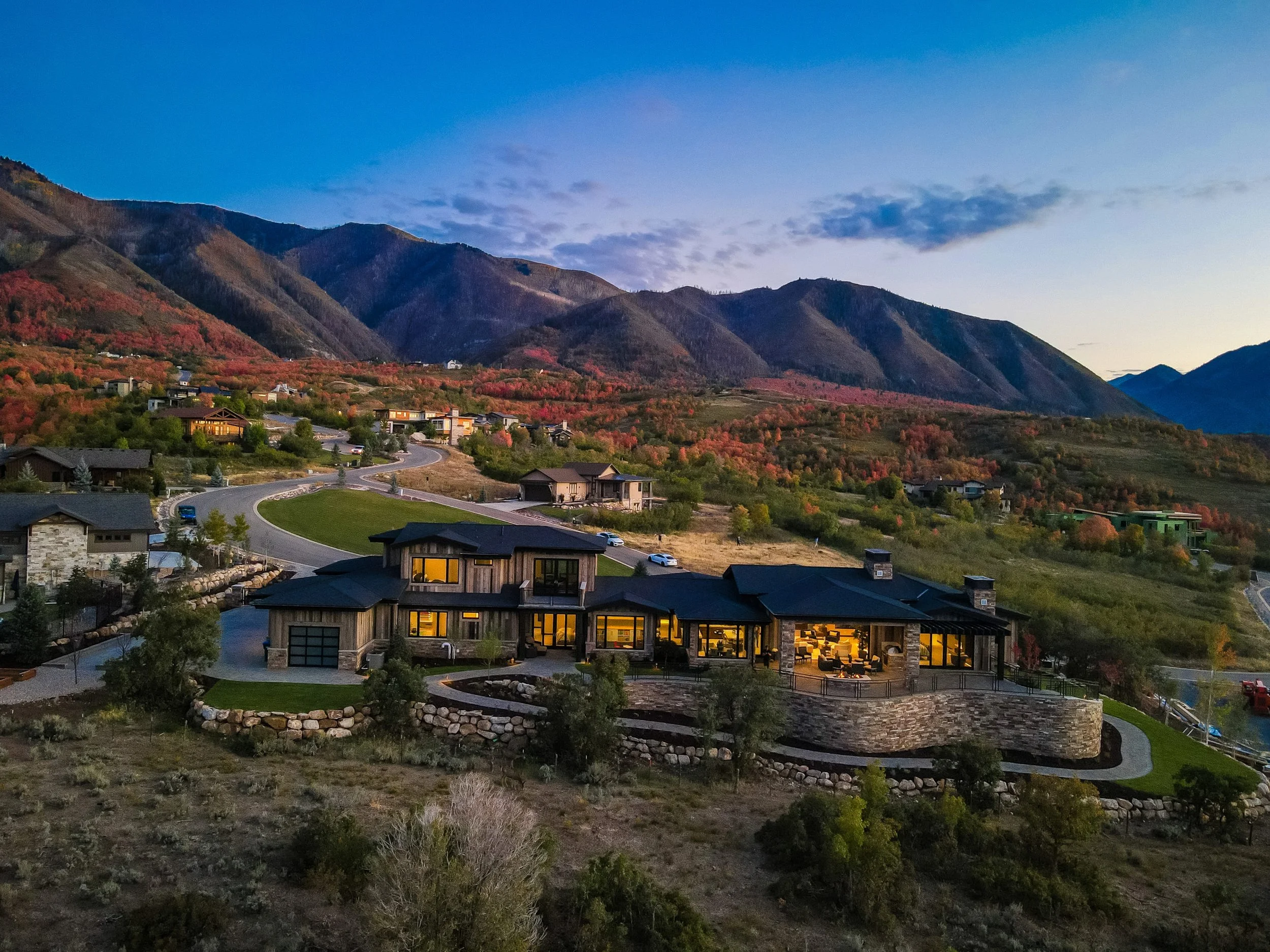 An aerial view of a modern house with large windows, illuminated from inside, situated on a hillside with a winding road. In the background are mountains and trees with fall foliage, under a partly cloudy sky at dusk.