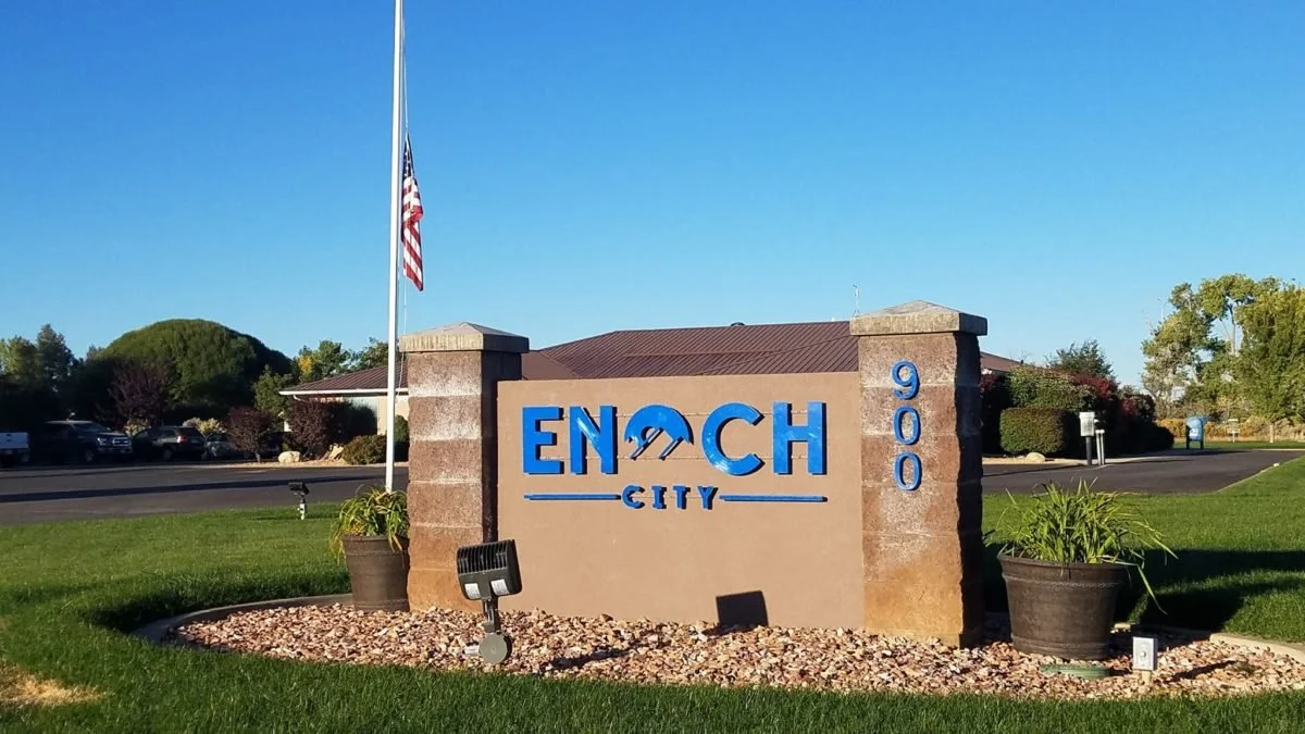 Entrance sign for Enoch City with a blue and beige stone design, an American flag on a flagpole, and a landscaped area with potted plants.