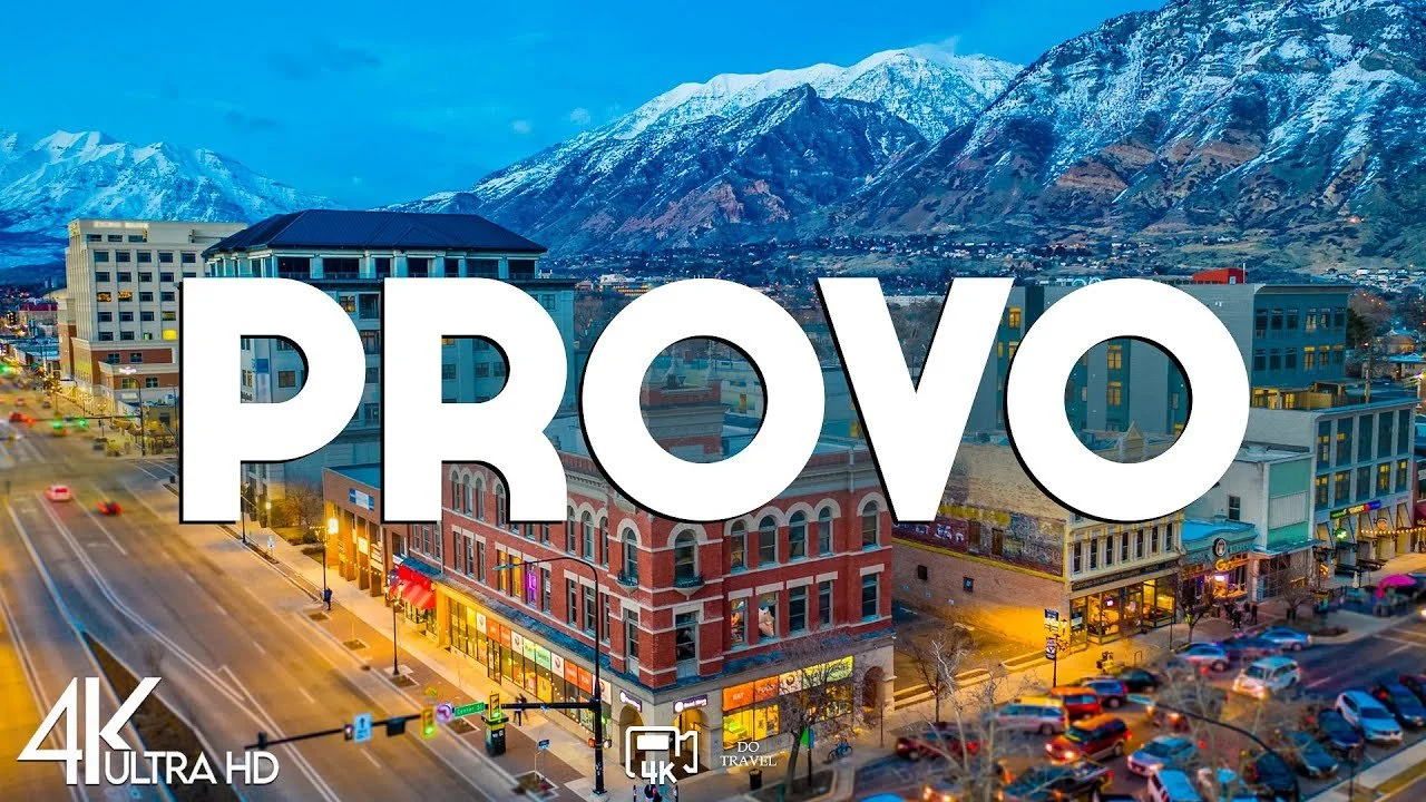 Downtown city street with colorful buildings, cars, and mountains in the background, overlaid with the word 'PROVO' in large white letters.