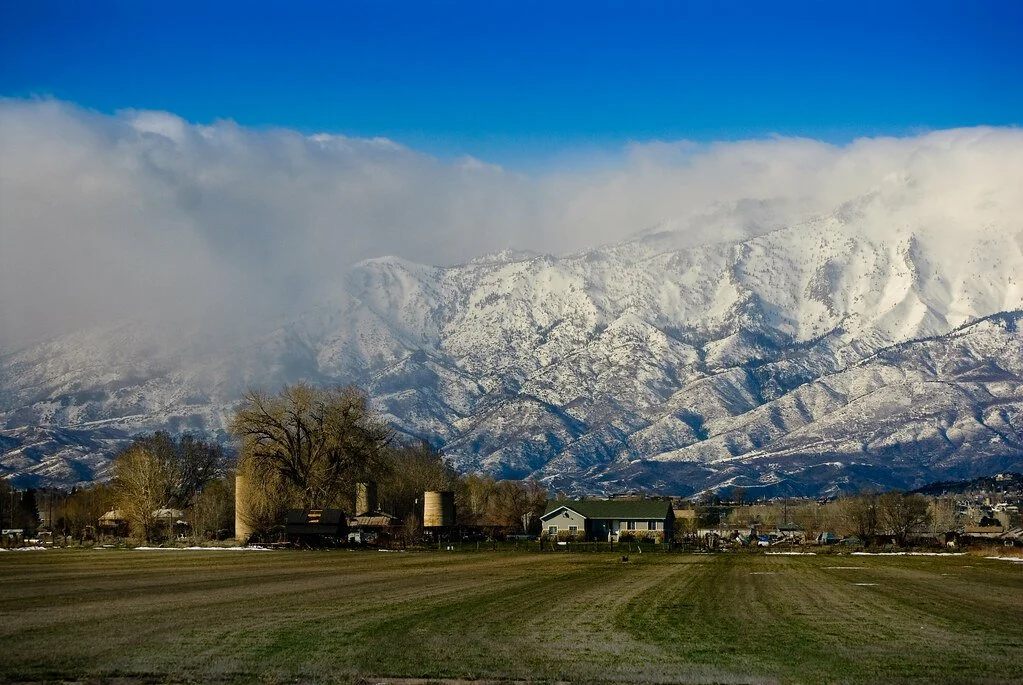 Snow-capped mountains with a small rural farm in the foreground and a clear blue sky with some clouds.