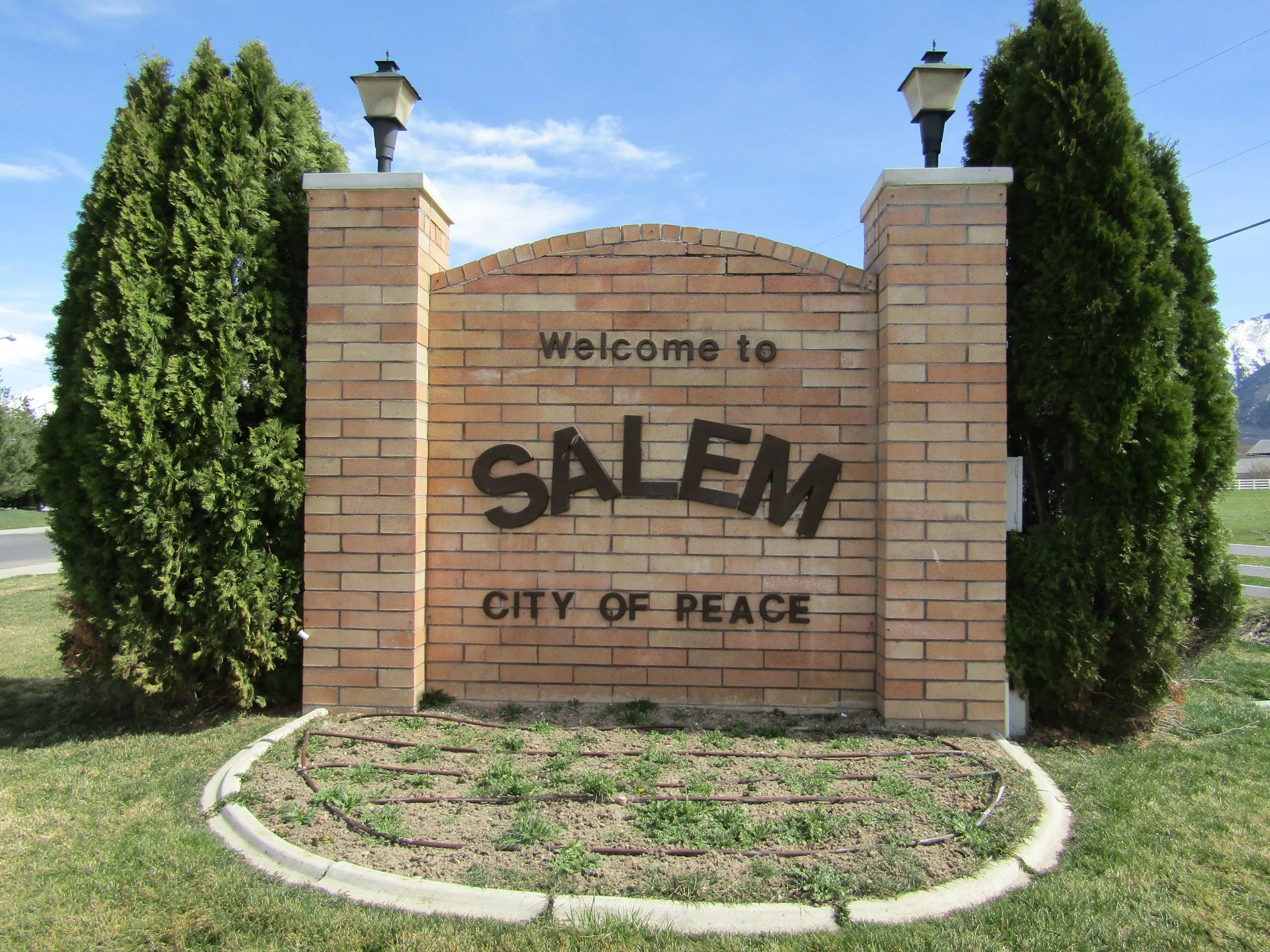Welcome to Salem, City of Peace sign on brick wall, flanked by two tall green trees, with grass and a small garden bed at base, under a blue sky.